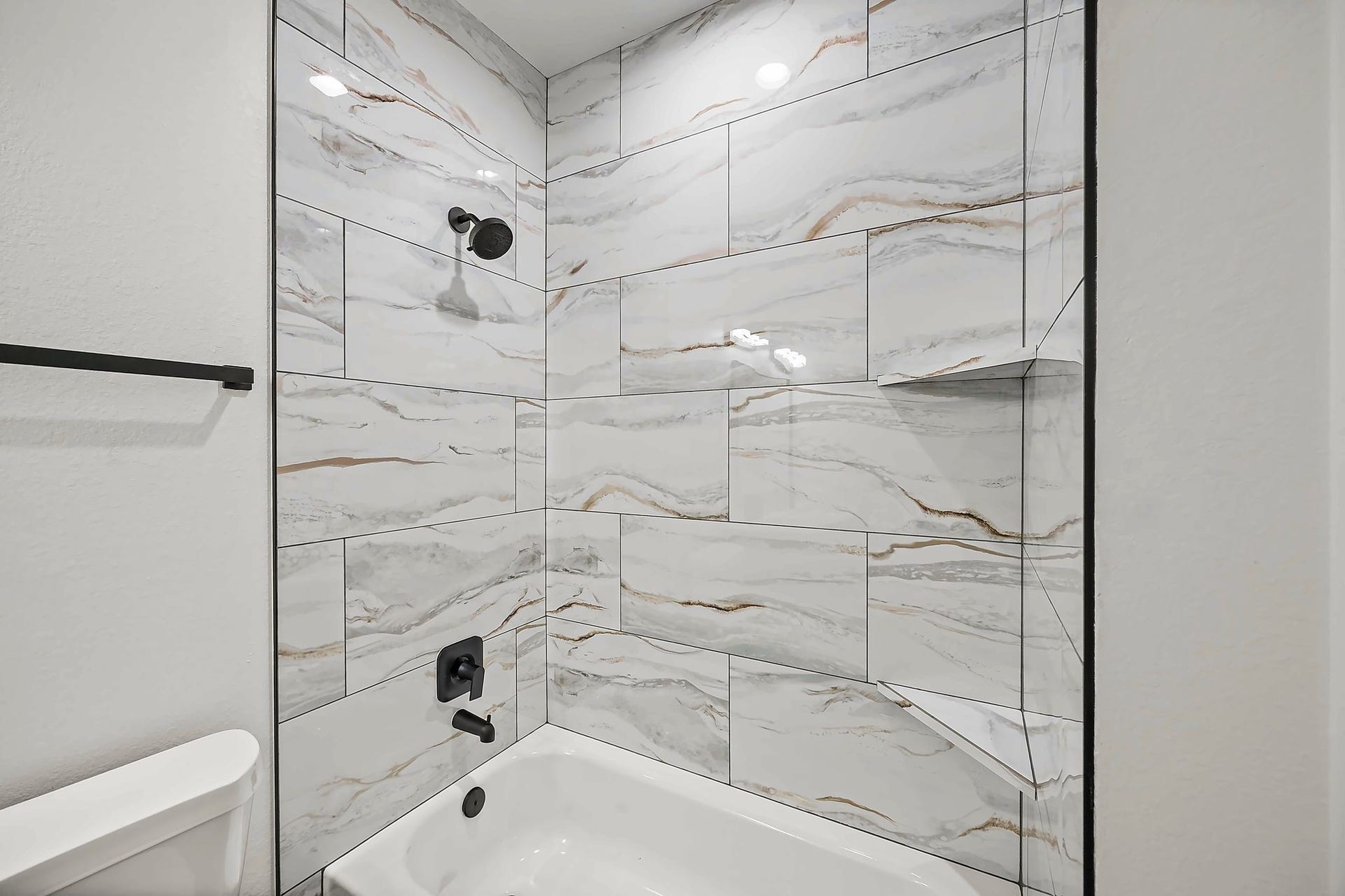 10426 Royal Oaks Drive, Houston, TX 77016 Luxurious bathroom featuring elegant marble-style tilework, a sleek black shower fixture, and a pristine white bathtub for a modern touch.