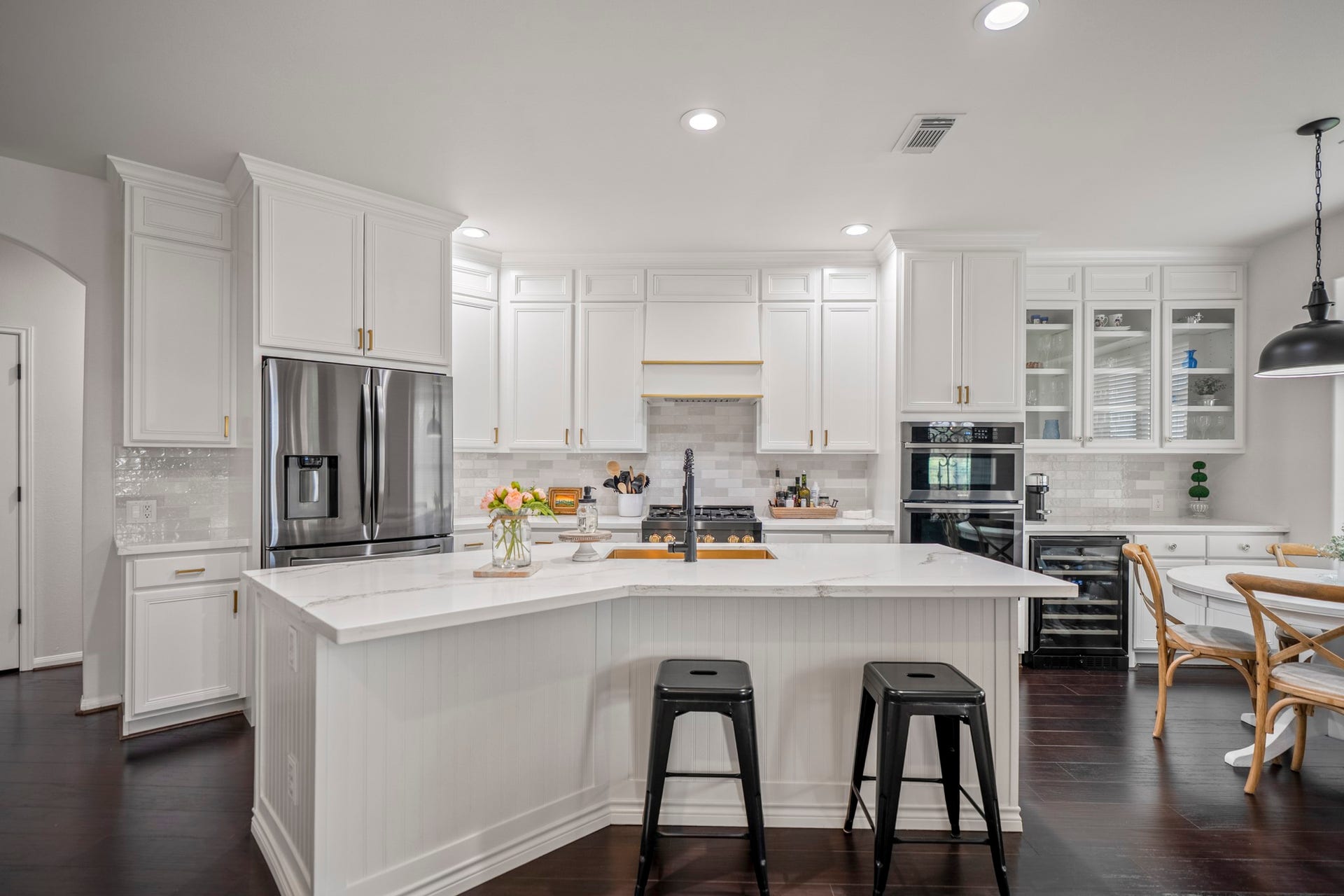 Crisply painted white cabinetry extends to the ceiling, perfectly accented by brushed gold hardware, glass fronted display cabinetry above a beverage station, and modern tile backsplash, with custom vent hood cover.