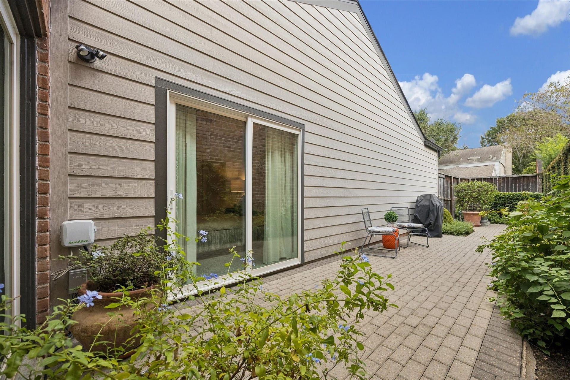 Spacious brick-paved side patio offers an inviting outdoor entertaining area surrounded by greenery, with convenient access to the home.