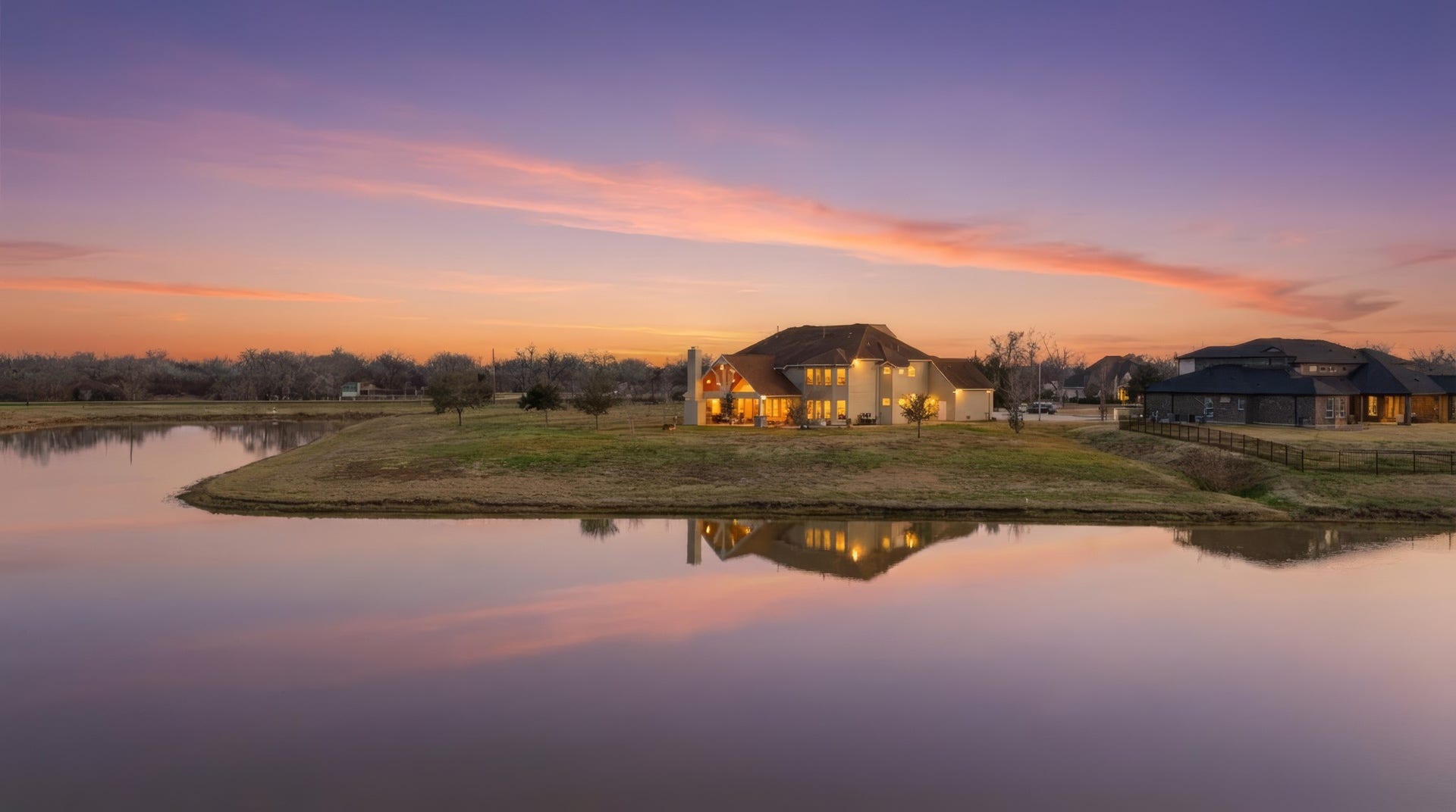 As the sun sets in a wash of soft blush and lavender tones, this exceptional waterfront estate glows warmly against the horizon. Perfectly positioned along the waters edge, the homes reflection shimmers across the tranquil pond, creating a breathtaking evening backdrop that feels both private and resort-inspired.