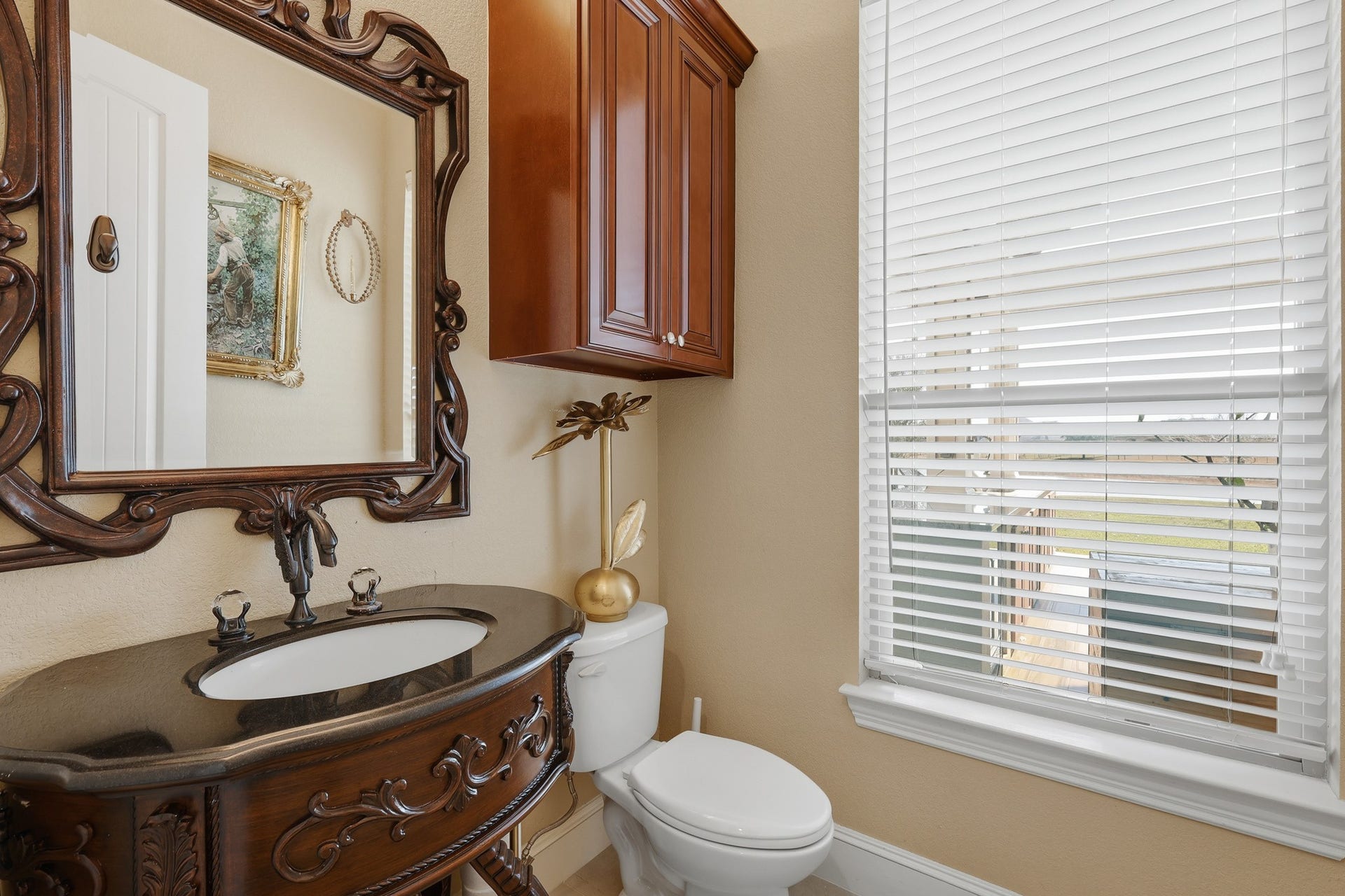 Conveniently located just off the main living, this elegant first-floor powder bath makes a refined statement. A richly detailed custom wood vanity with a sculpted base is topped with a sleek stone countertop, while an ornate framed mirror adds timeless sophistication. Warm finishes, designer fixtures, and natural light from the window create an inviting yet luxurious spaceperfectly positioned for guests and everyday convenience.