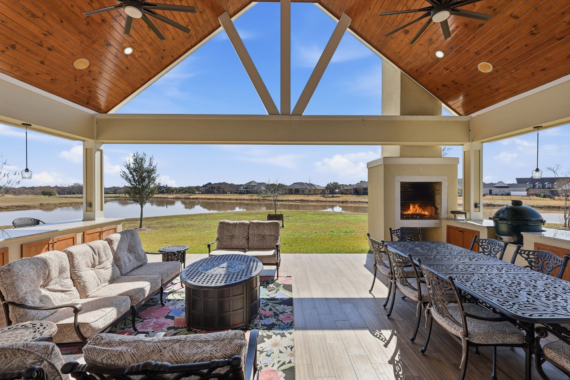 Designed for year-round entertaining, this expansive covered patio showcases soaring wood-paneled ceilings, statement ceiling fans, and panoramic water views. A stunning outdoor fireplace anchors the space, while a fully equipped outdoor kitchen and elegant dining area create the ultimate setting for refined al fresco gatherings.