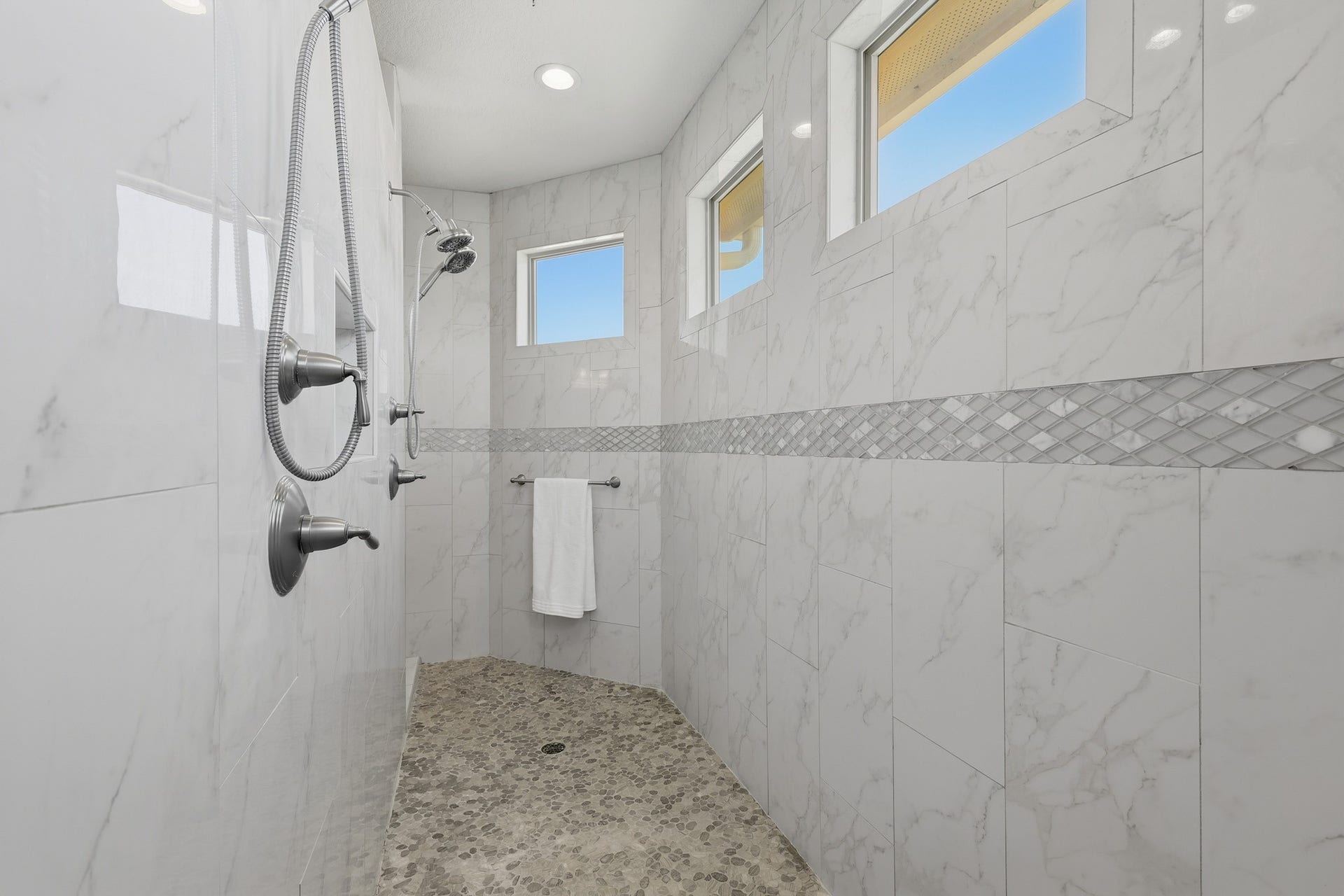 The second-floor primary suite showcases a stunning walk-in shower wrapped in floor-to-ceiling marble-look tile, accented with decorative mosaic detailing and a pebble stone floor. Multiple windows invite natural light, enhancing the clean lines and luxurious spa-like ambiance of this expansive shower space.