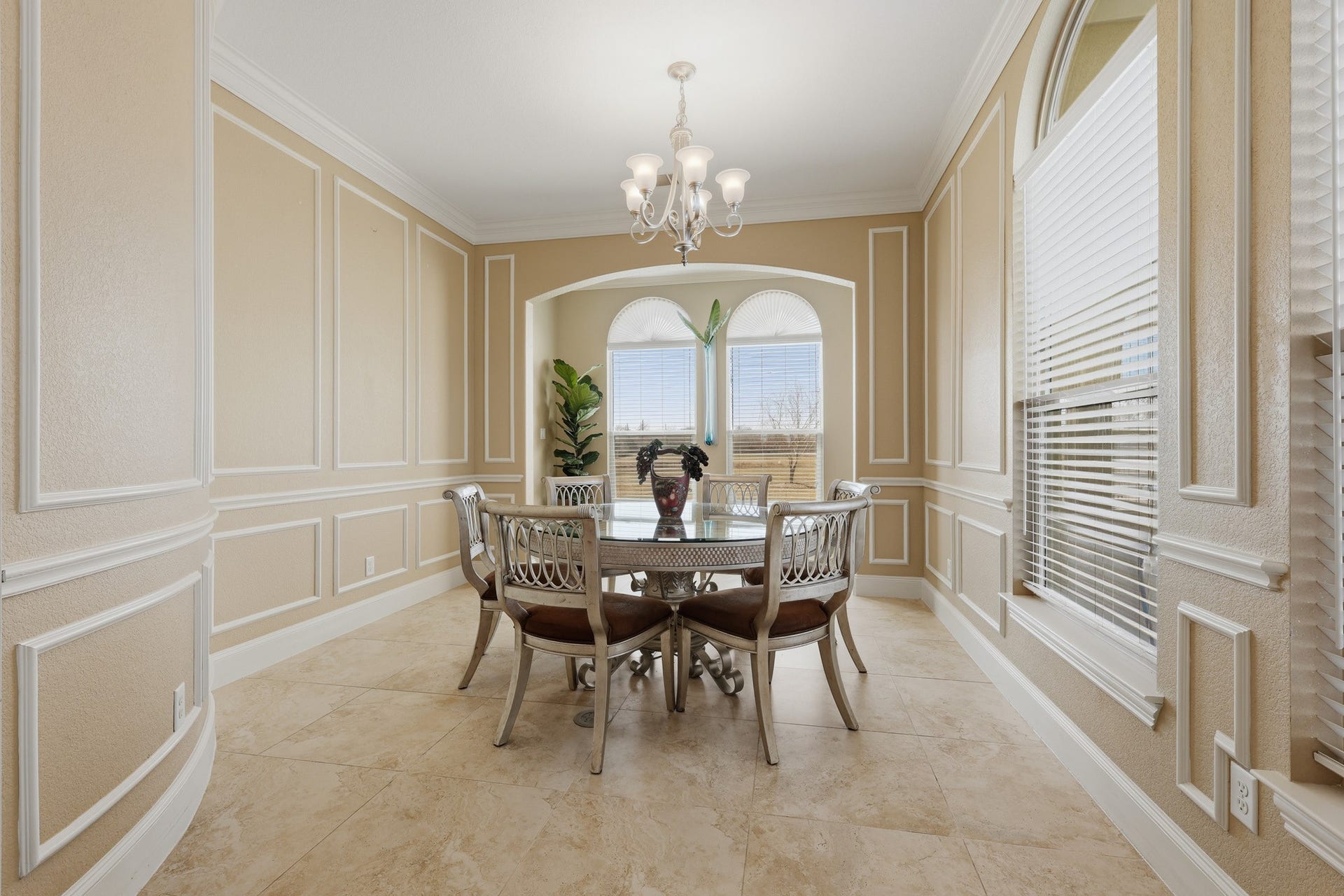 This formal dining room exudes timeless sophistication. Framed by custom wall panel molding and crowned with classic millwork, the space offers architectural depth and distinction throughout. Rich stone flooring enhances the rooms upscale appeal, while the arched alcove with twin windows draws in beautiful natural light and frames serene outdoor views. neutral palette provides a versatile backdrop allowing the space to transition effortlessly from formal celebrations to elevated everyday dining.