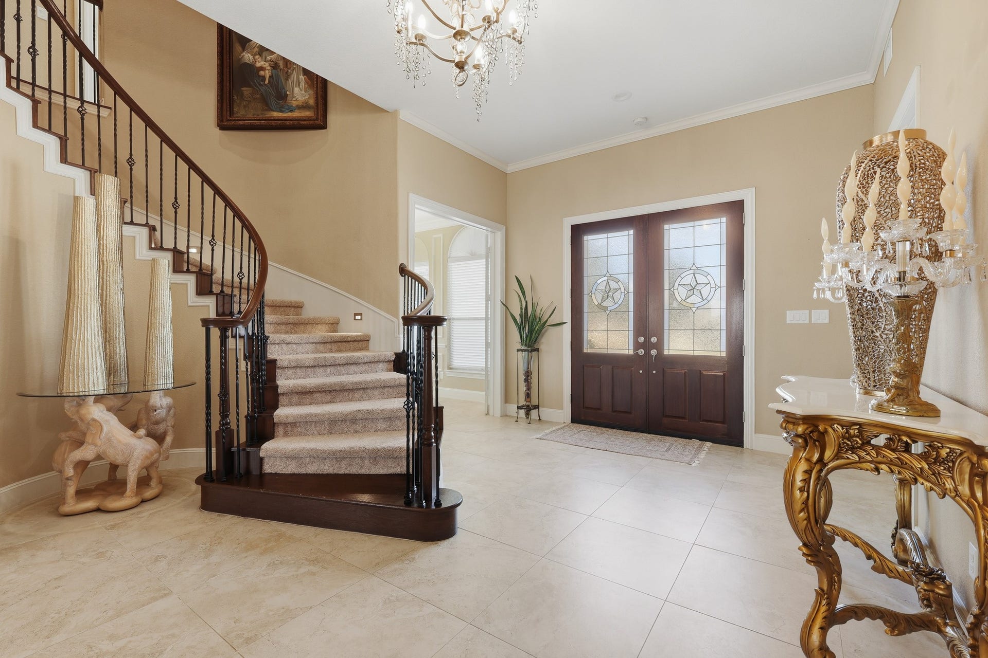 A breathtaking first impression awaits in this elegant, gallery-inspired foyer. Soaring ceilings and a sweeping curved staircase with rich wood treads and custom iron balusters create an architectural statement of timeless sophistication. The double front doors, adorned with decorative glass inserts, invite natural light to pour across the polished stone flooring, enhancing the warm, neutral palette.