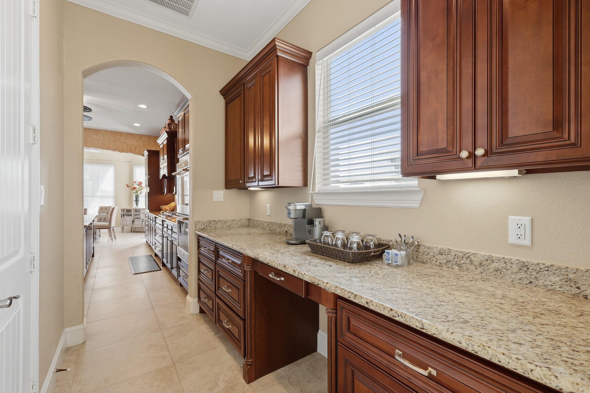 A beautifully appointed butlers pantry offering exceptional versatility and elegance. Rich custom cabinetry in a warm cherry finish pairs seamlessly with polished granite countertops, creating an ideal space for coffee service, entertaining prep, or additional storage. Natural light streams through the window, enhancing the refined details and thoughtful functionality of this sophisticated extension of the kitchen.