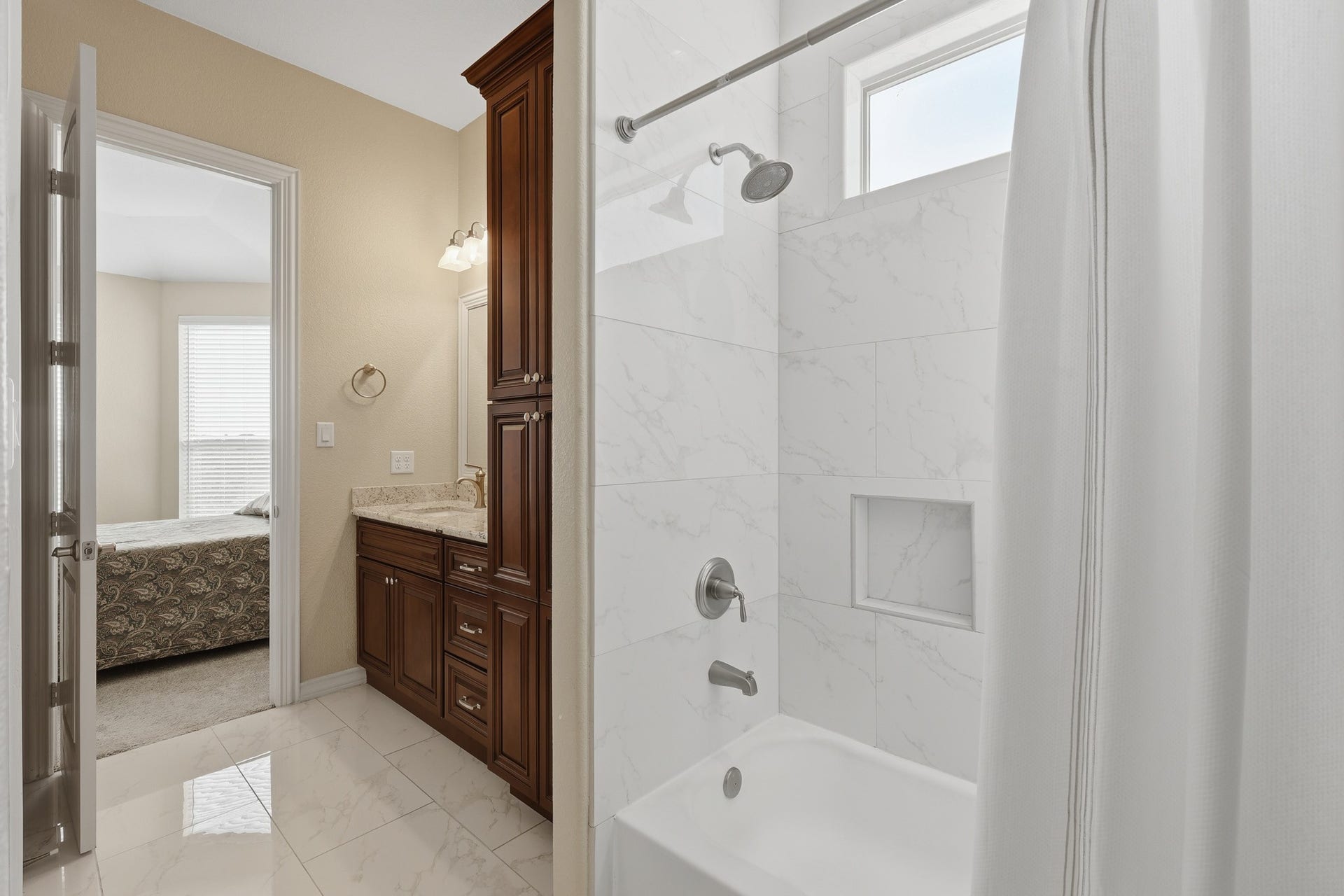 This private en suite bath showcases custom cherry wood cabinetry paired with granite countertops, creating a warm and sophisticated contrast to the light, marble-inspired tile that surrounds the tub and shower. Polished fixtures and a built-in shower niche enhance the clean, refined aesthetic, while natural light brightens the space. Seamlessly connected to the bedroom, this well-appointed en suite also features a large walk-in closet, offering generous storage and organization.