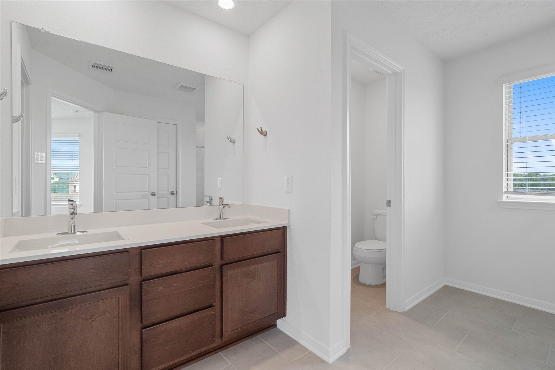 The primary bathroom is move-in ready! It showcases stained cabinetry paired with light countertops, dual vanity sinks, a large mirror, a spacious walk-in closet with built-in shelving, toilet compartment, high ceilings, custom paint, and stylish modern finishes throughout.