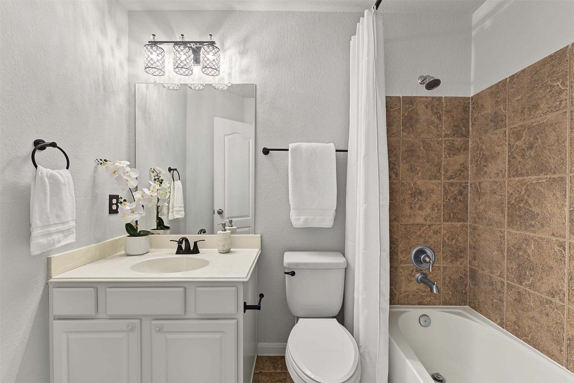 Charming secondary bathroom with elegant lighting, a sleek vanity, and a tub featuring warm tiled walls for a cozy retreat.