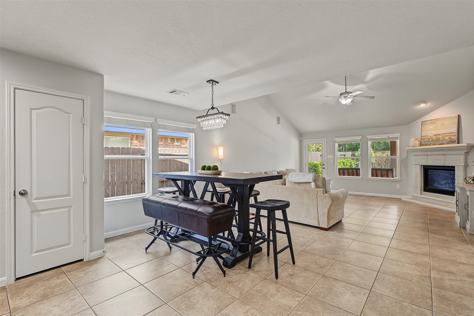 Bright and spacious open-plan living area featuring a cozy fireplace, modern chandelier, and ample natural light from large windows overlooking the backyard.