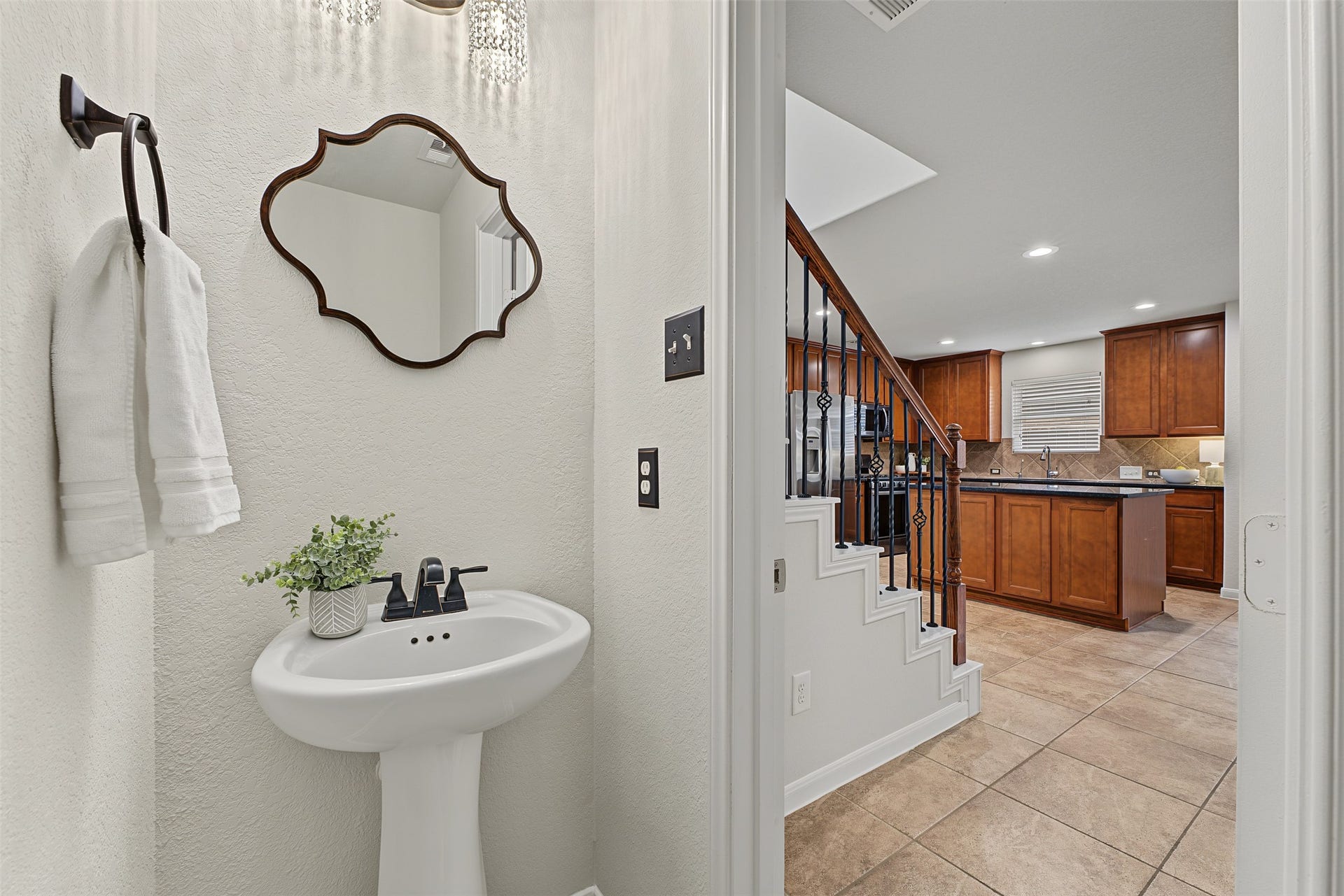 A stylish powder room with a pedestal sink and decorative mirror, leading to a modern kitchen with rich wood cabinetry and sleek countertops.