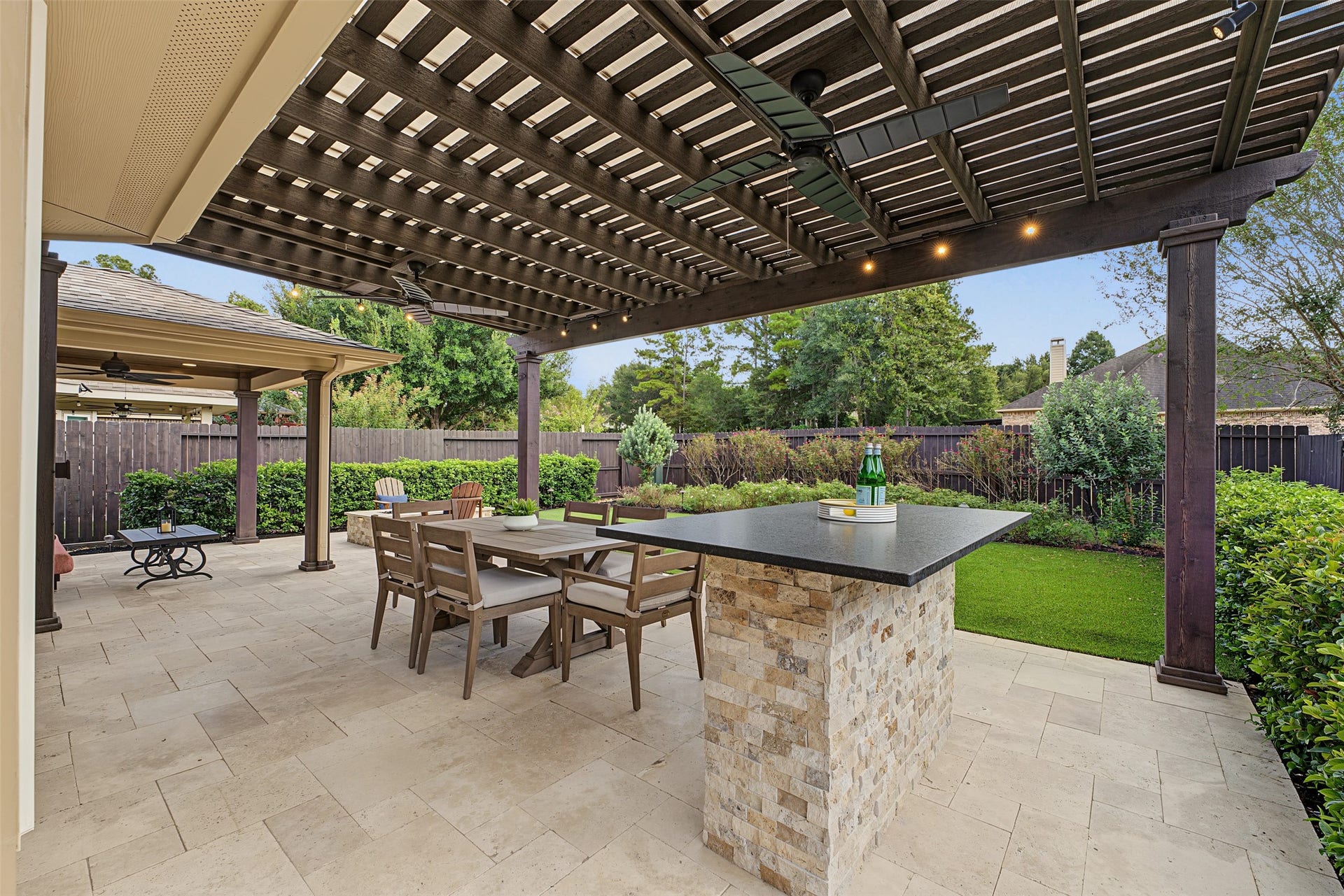 Experience outdoor living at its finest with this elegant patio featuring a stylish pergola, a spacious dining area, and a sleek stone bar, all set against a lush, manicured garden backdrop and artificial turf.