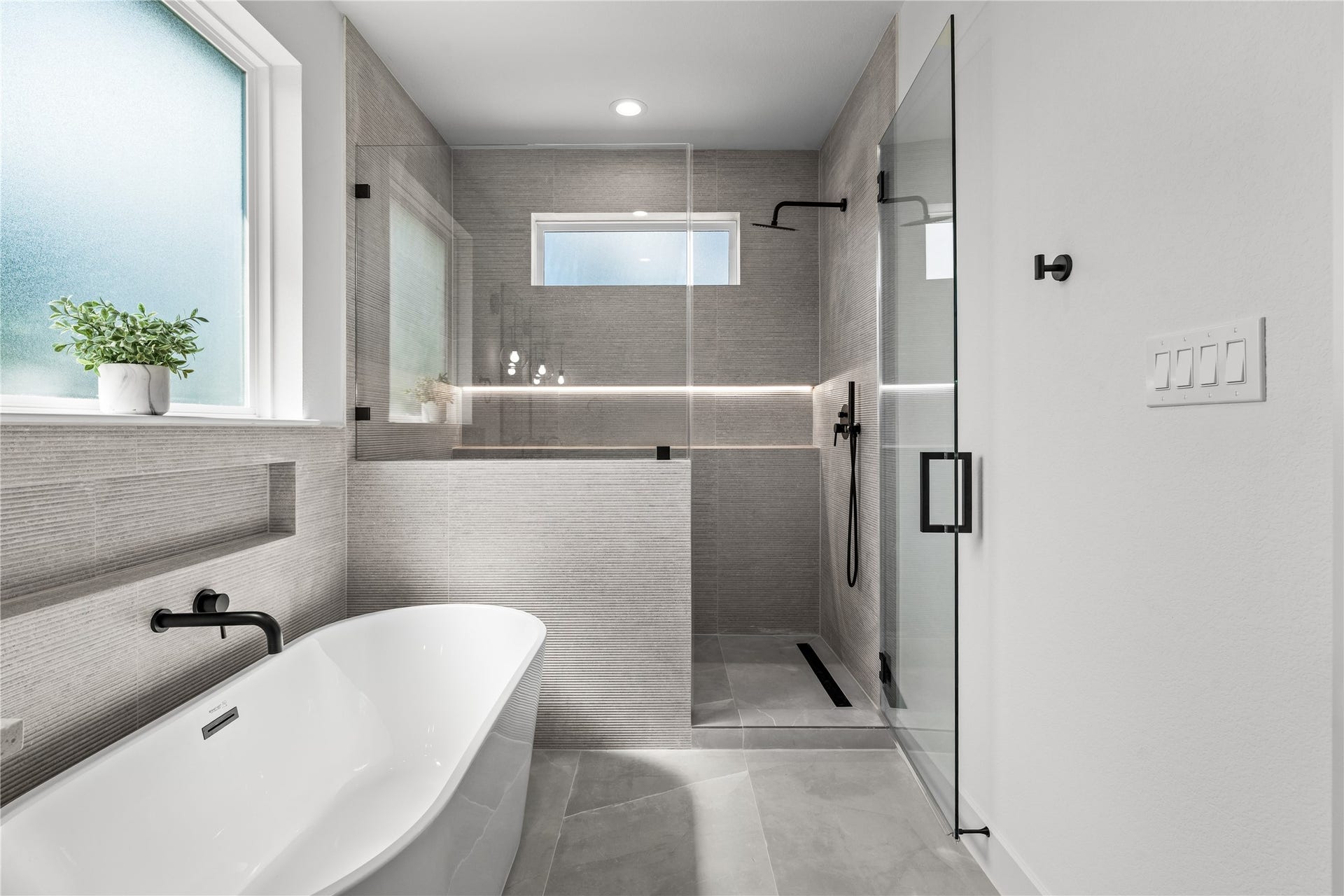 3012 & 3014 Beulah St, Houston, TX 77004 The primary shower features a seamless glass enclosure and neutral floor-to-ceiling tiling. It includes a modern black rainfall showerhead, a built-in tile niche, and a linear drain, sitting right next to the freestanding soaking tub under a large frosted window to create a clean, spa-like wet room.