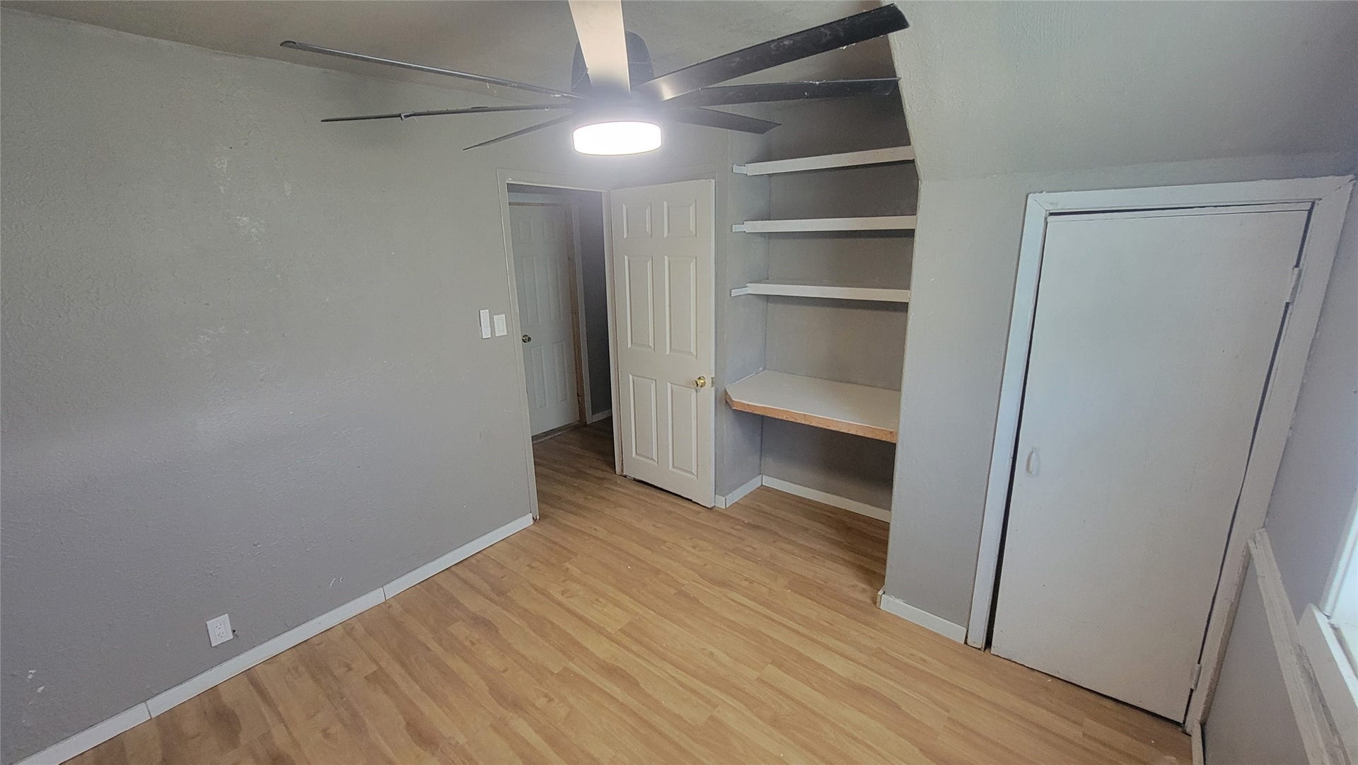 12018 Sandpiper Road, Angleton, TX 77515 This cozy room features laminate wood flooring, gray walls, a ceiling fan with light, built-in shelves, and a small closet. The door leads to a hallway, and a window allows natural light to brighten the space. Ideal for a small bedroom or office.