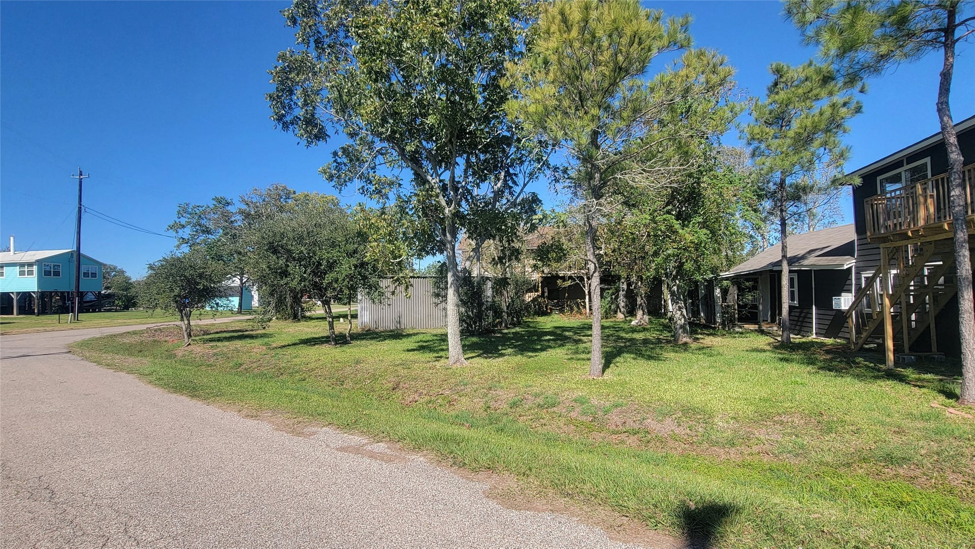 12018 Sandpiper Road, Angleton, TX 77515 This photo shows a quiet residential street with a grassy lot and mature trees. Nearby, there are raised homes with wooden decks, suggesting a coastal or flood-prone area. The setting appears peaceful and spacious.