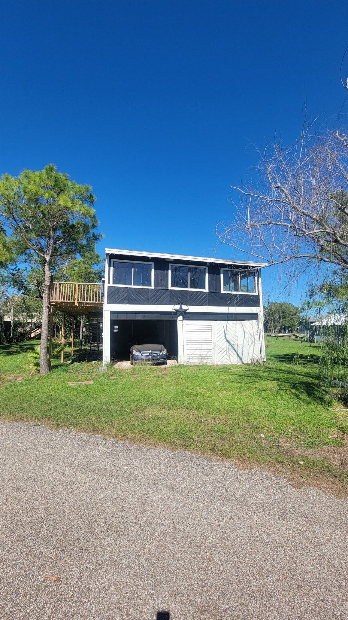 12018 Sandpiper Road, Angleton, TX 77515 This is a two-story home with a screened upper level and a covered carport below. It has a driveway leading up to the carport and is surrounded by trees, set against a clear blue sky.