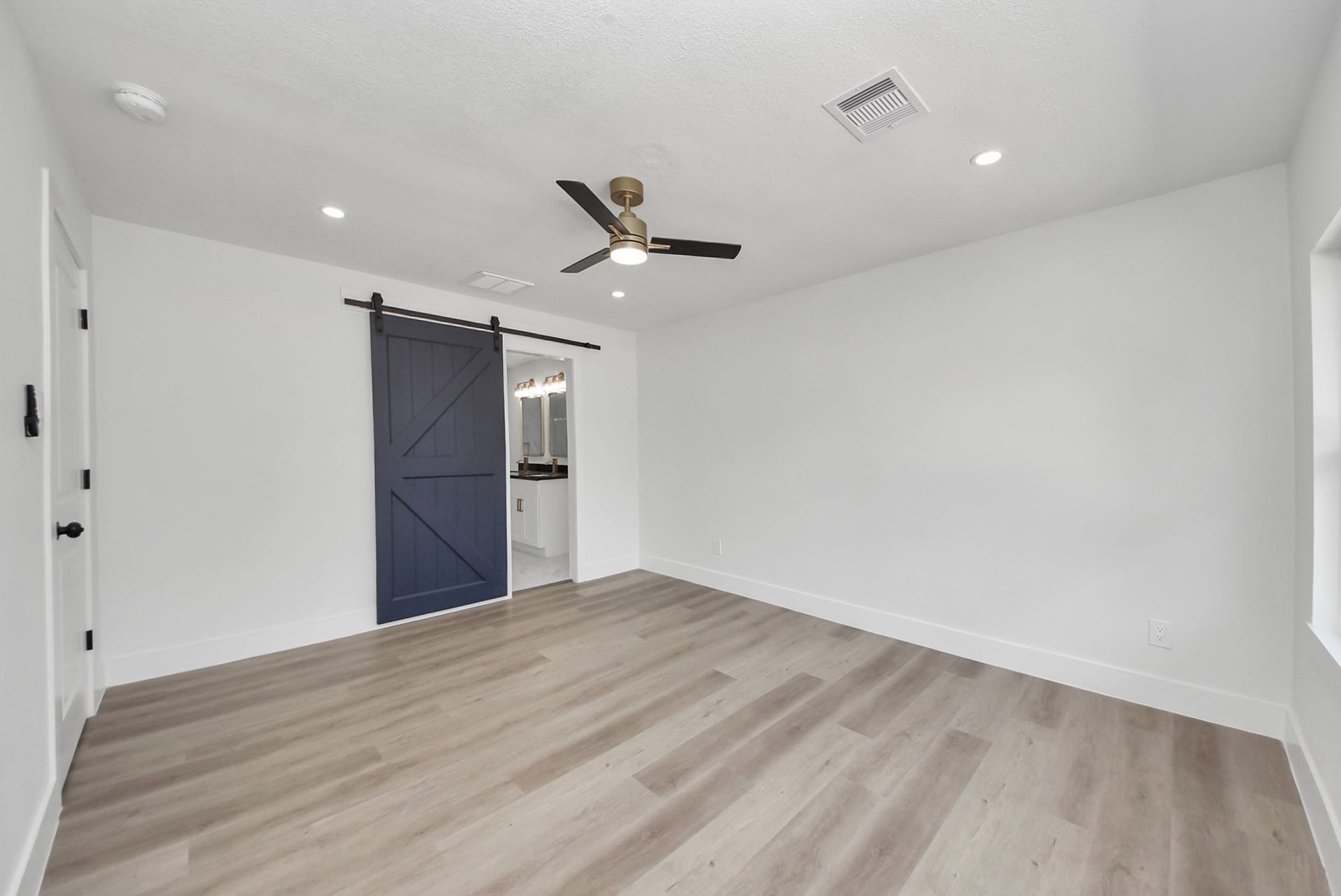 602 Gammon Drive, Houston, TX 77022 Spacious room with elegant wood flooring, modern recessed lighting, and a stylish ceiling fan, featuring a striking blue barn door leading to a well-lit bathroom.