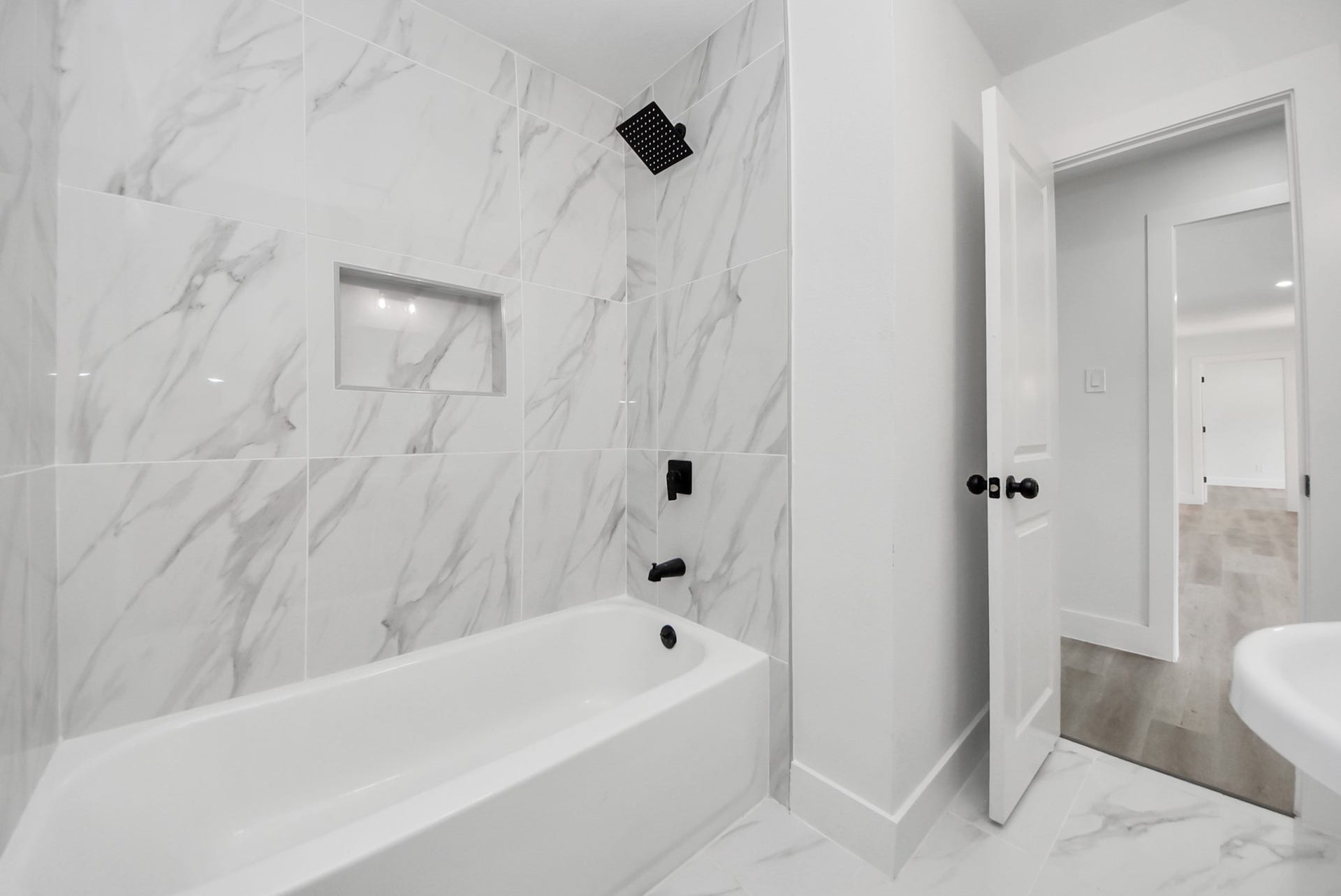 602 Gammon Drive, Houston, TX 77022 Sleek bathroom featuring elegant marble-patterned tiles, a modern black shower fixture, and a convenient built-in shelf above a pristine bathtub.