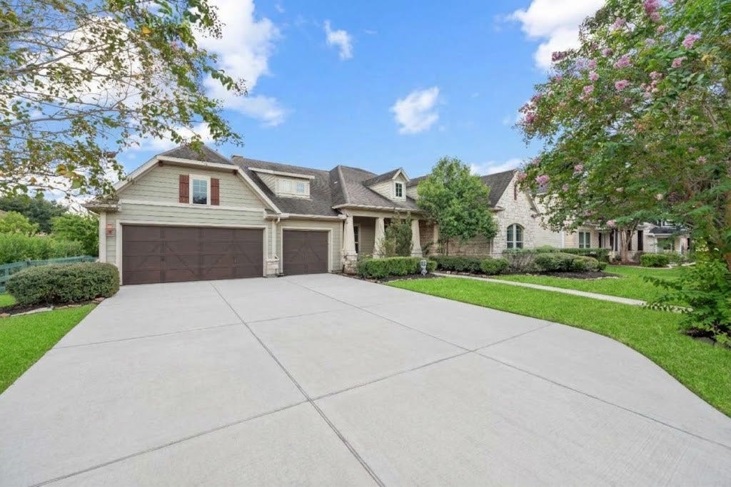 5414 Lake Hill Farm Way, Fulshear, TX 77441 Driveway view highlighting the attached multi-car garage, stone and lap-siding exterior, covered entry porch, and landscaped front lawn with stone-edged flower beds.