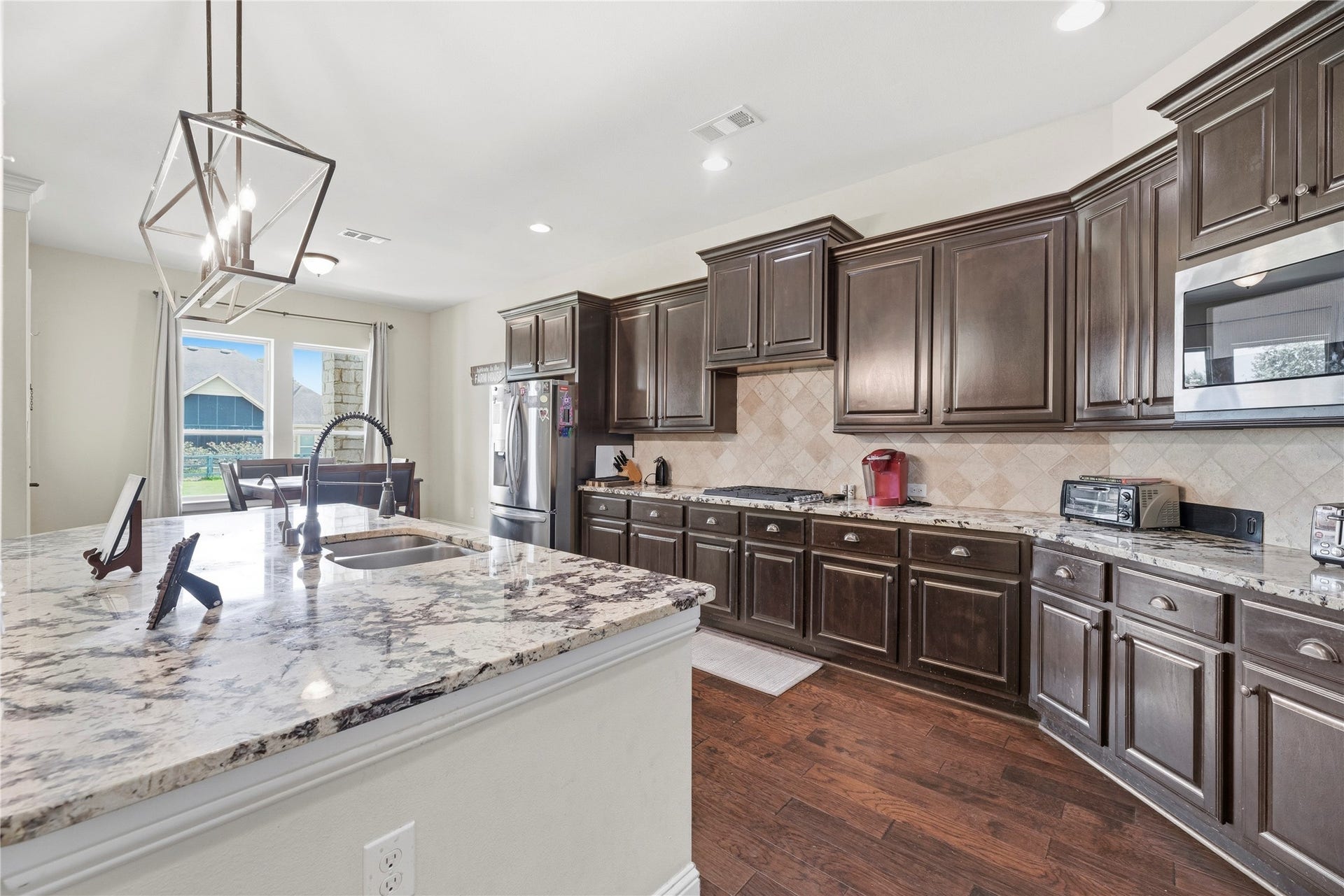 5414 Lake Hill Farm Way, Fulshear, TX 77441 Spacious kitchen showcasing rich dark-stained cabinetry, granite countertops, stone tile backsplash, a multi-seat island with an undermount sink and gooseneck faucet, stainless steel appliances, and dark wood-look flooring.