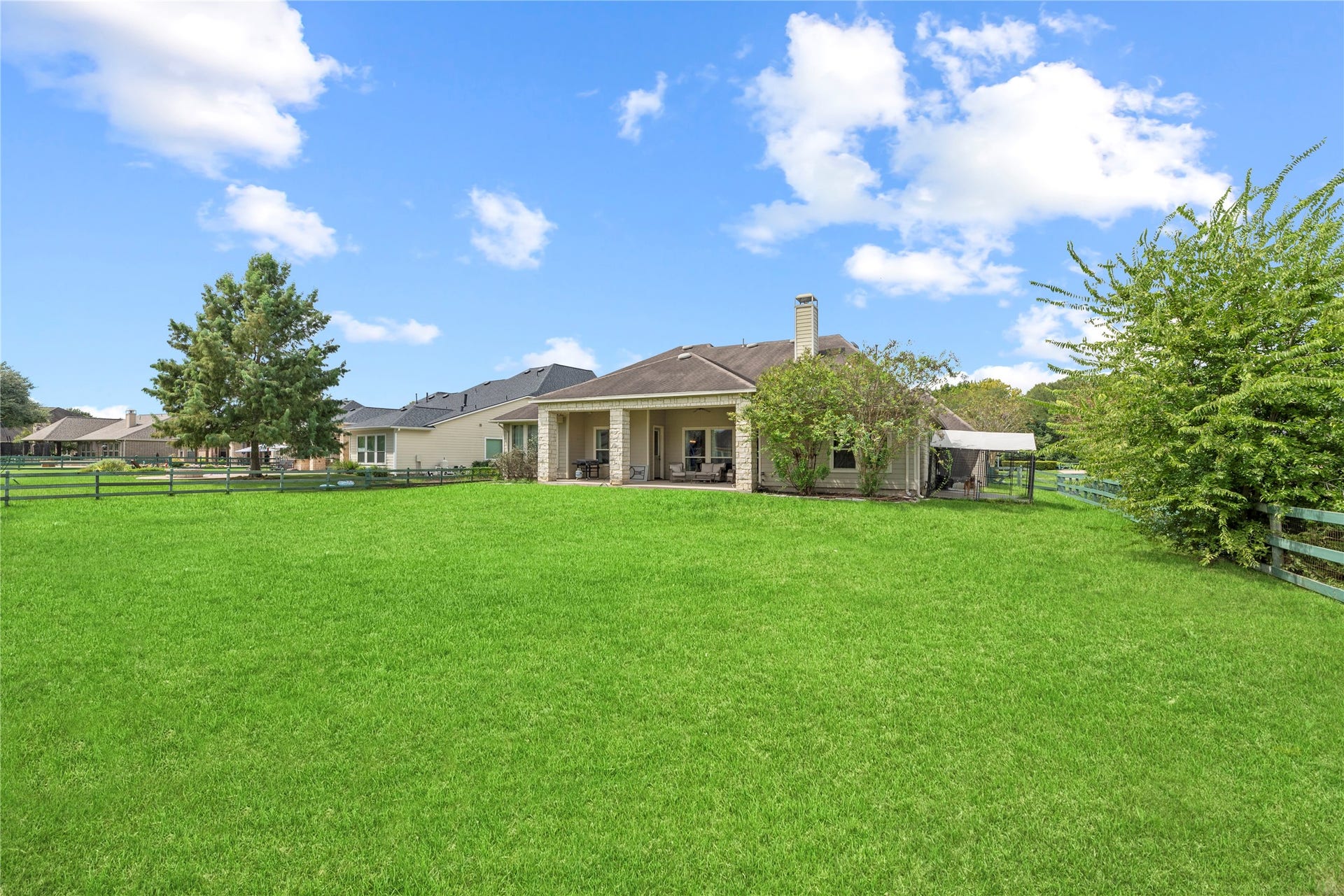 5414 Lake Hill Farm Way, Fulshear, TX 77441 Sprawling rear lawn highlighting the open green space, covered back porch with stone support pillars, exterior siding and stone trim, and bounding wood-rail fencing.