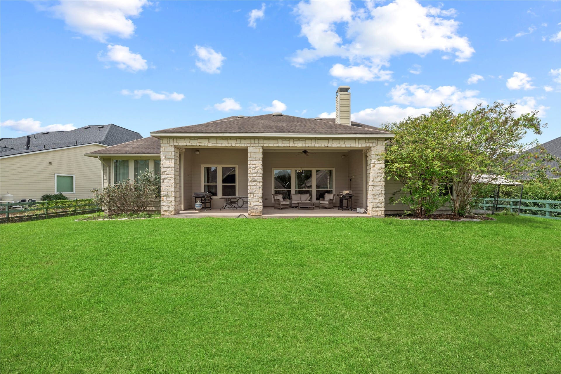 5414 Lake Hill Farm Way, Fulshear, TX 77441 Centered view of the covered outdoor back patio framed by heavy stone pillars, featuring wide glass windows and doors along the rear wall, a roofline chimney, and a flat grass backyard.