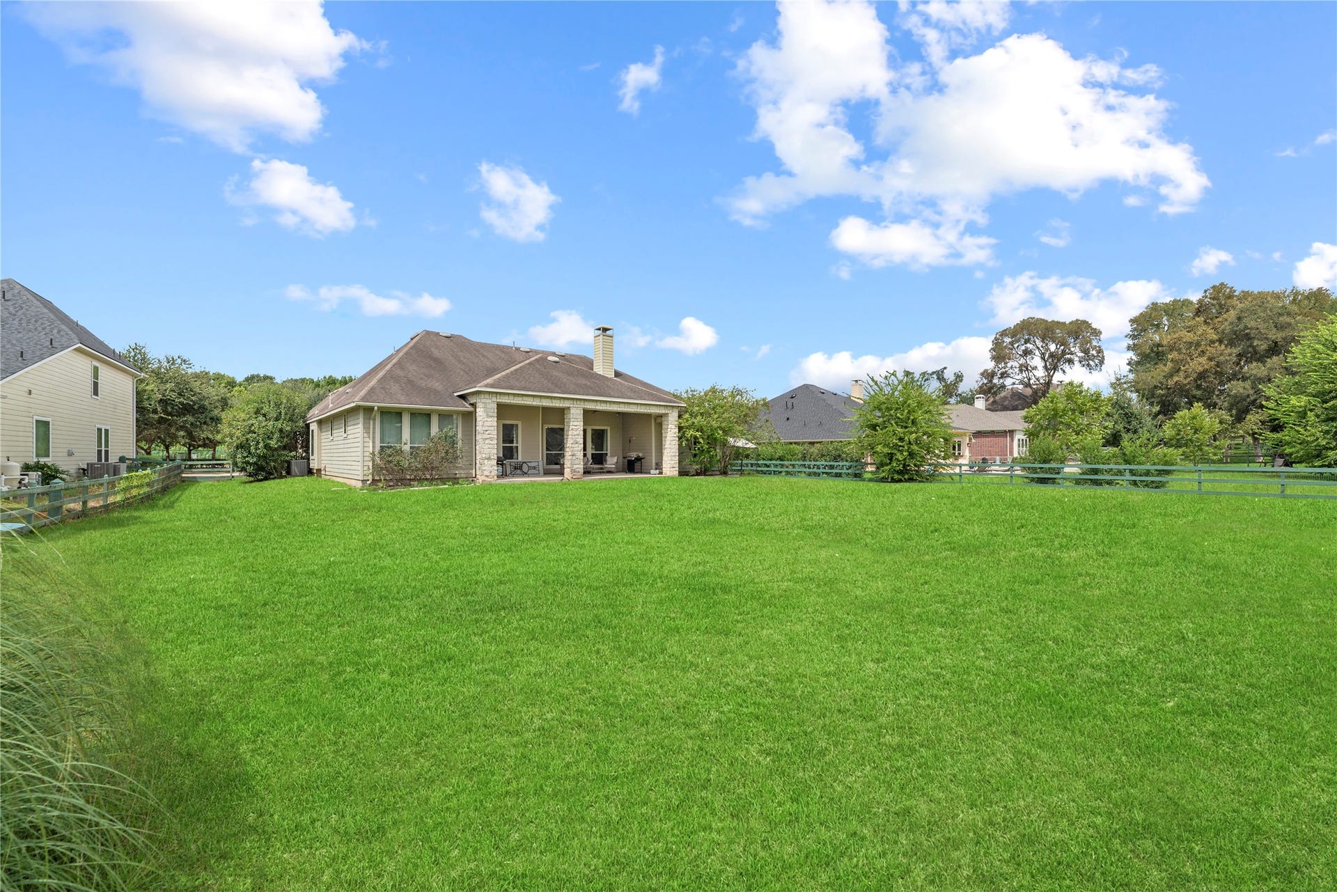 5414 Lake Hill Farm Way, Fulshear, TX 77441 Wide-angle view of the expansive, level grass yard showcasing the stone-trimmed rear elevation, covered patio with supporting stone pillars, hip roofline, exterior chimney stack, and green perimeter post-and-rail fencing.