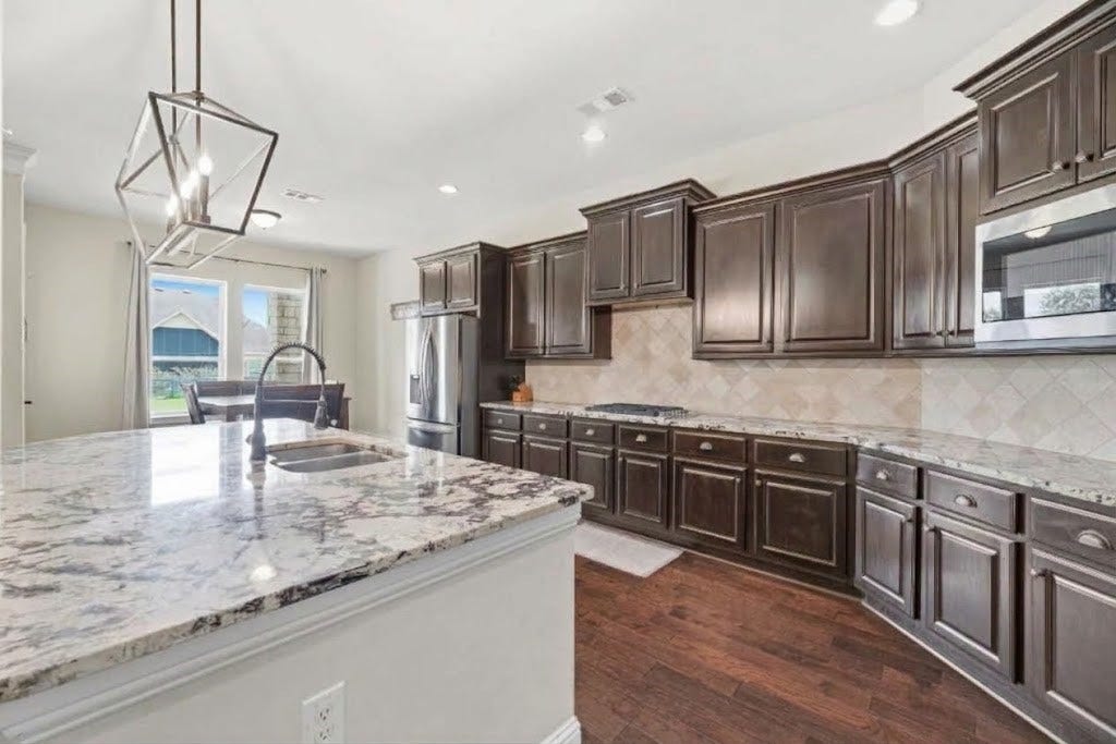 5414 Lake Hill Farm Way, Fulshear, TX 77441 Angle focusing on the large granite island breakfast bar with hanging lantern light fixture, deep undermount sink, gooseneck faucet, dark stain cabinetry, and seamless connection to the dining nook.