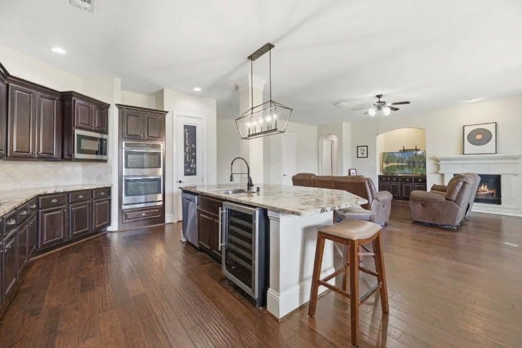 5414 Lake Hill Farm Way, Fulshear, TX 77441 Wide view of the open kitchen and living area, showcasing the built-in wine cooler in the island, stainless steel appliances, dark wood-look flooring, and connected sightlines to the fireplace and living spaces.