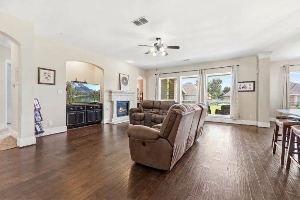 5414 Lake Hill Farm Way, Fulshear, TX 77441 Open-concept living room boasting rich dark wood-look flooring, high ceilings with a central ceiling fan, a corner gas-log fireplace with tile surround and mantle, built-in media nook, and a wall of large windows overlooking the backyard.