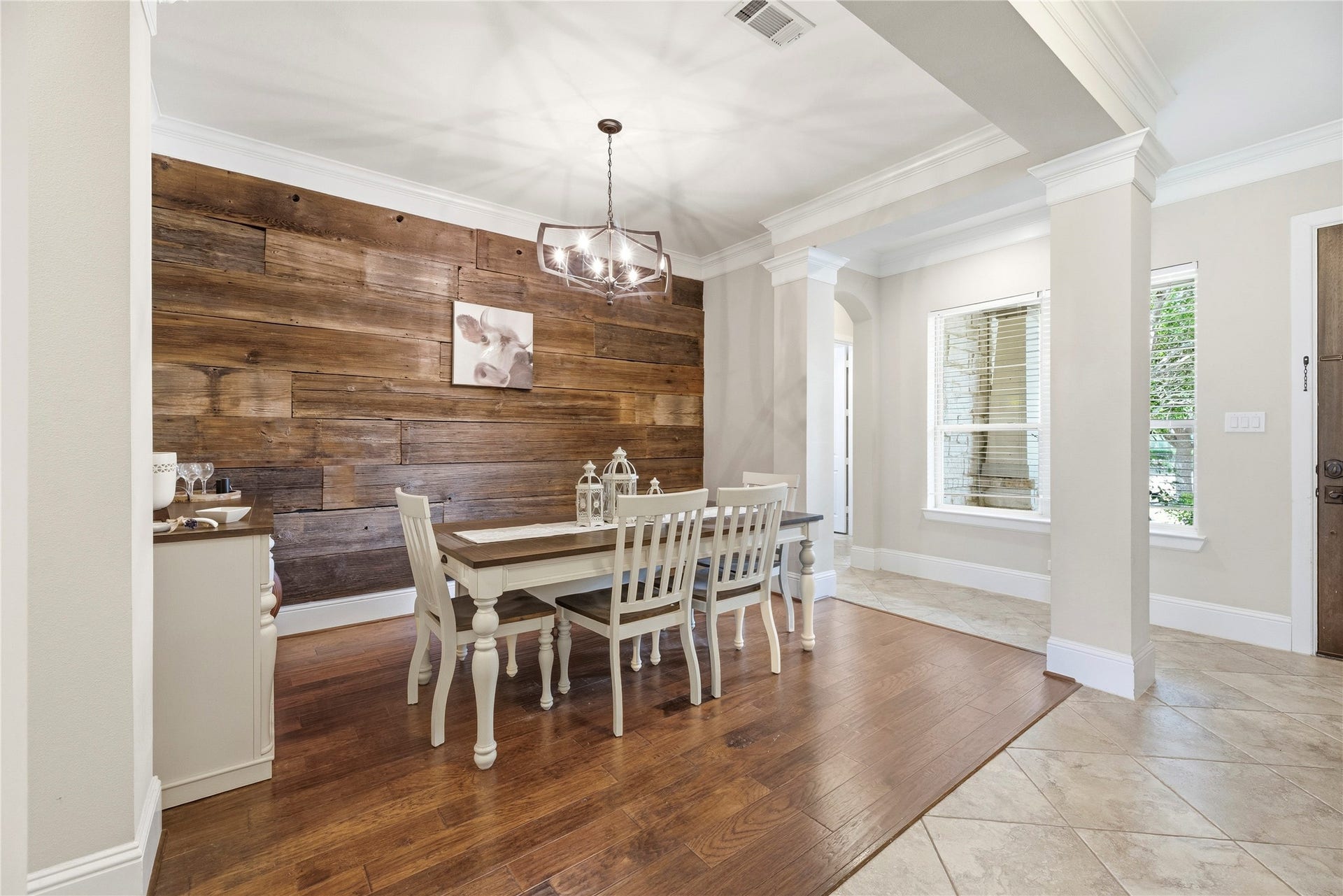 5414 Lake Hill Farm Way, Fulshear, TX 77441 Formal dining room with rich dark wood-look flooring, crown molding, an accent wood-plank wall, a modern hanging chandelier light fixture, and large side windows for natural light, framed by decorative drywall columns.