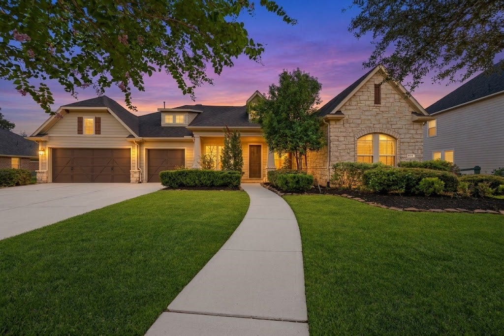 5414 Lake Hill Farm Way, Fulshear, TX 77441 Single-story home featuring a combination stone and horizontal siding facade, multi-gable roofline, front porch entry, three-car garage bay setup, concrete driveway and walkway, and mature professional landscaping with exterior accent lighting.