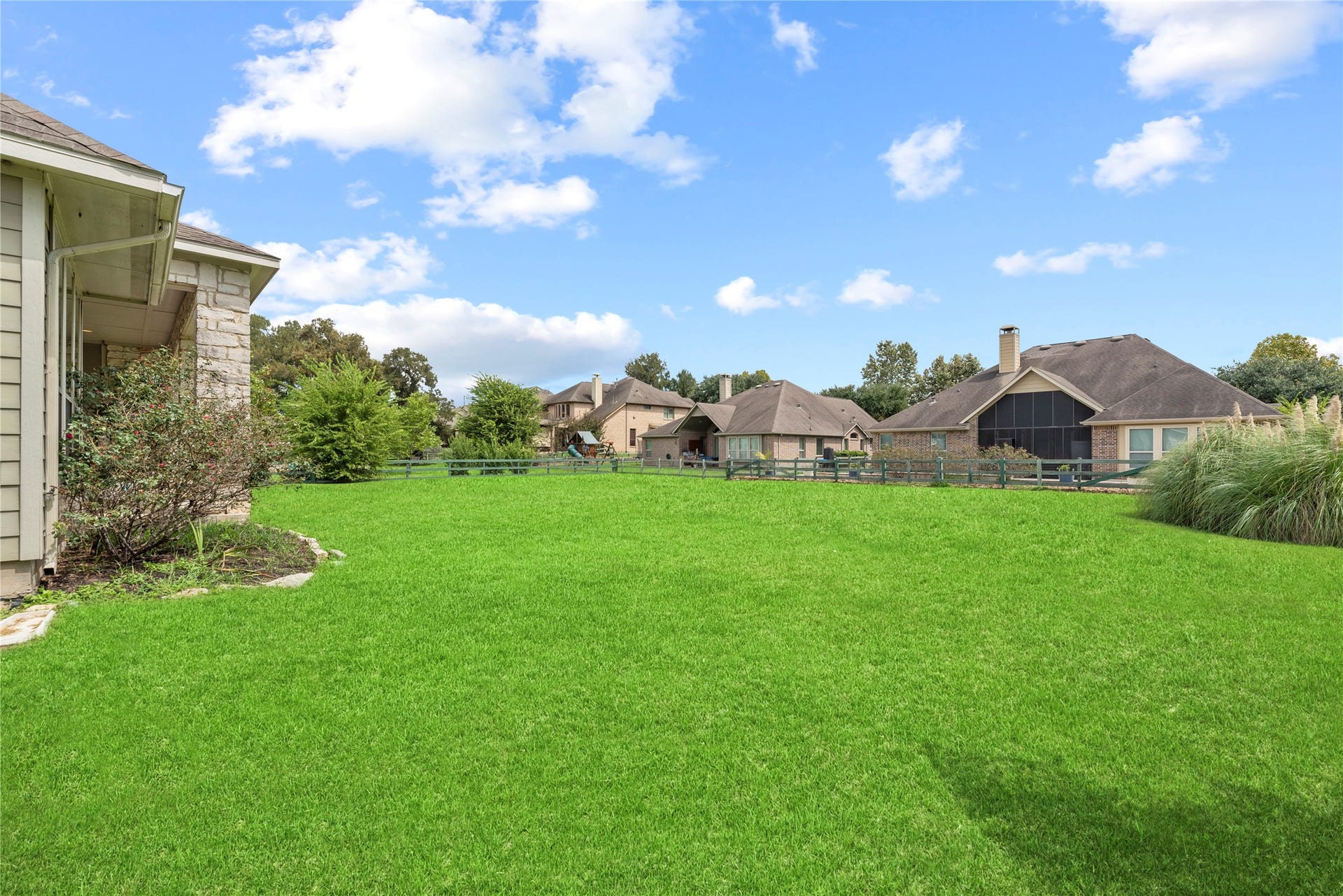5414 Lake Hill Farm Way, Fulshear, TX 77441 Angle view of the sprawling lawn and rear stone-and-siding exterior wall, highlighting the roofline, guttering, land slope, and perimeter wood-rail fencing.