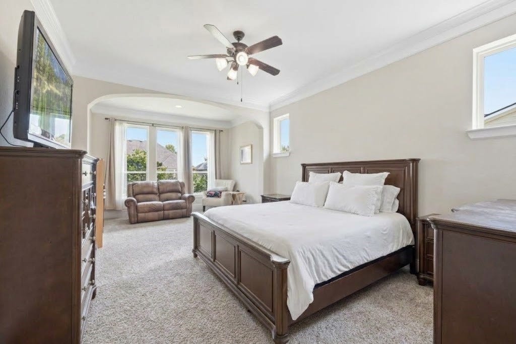 5414 Lake Hill Farm Way, Fulshear, TX 77441 Primary bedroom with plush wall-to-wall carpeting, high ceilings finished with crown molding, a central ceiling fan with light fixture, high accent windows, and a wide arched opening leading to an attached sitting nook/alcove.