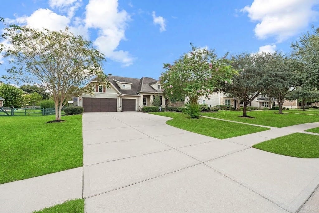 5414 Lake Hill Farm Way, Fulshear, TX 77441 Front perspective showing an expansive concrete driveway, three-car garage bay, combination stone and siding exterior facade, multi-gable roofline, and manicured front lawn with mature shade trees.