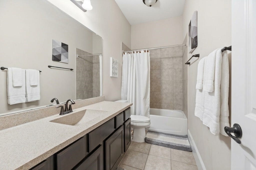 5414 Lake Hill Farm Way, Fulshear, TX 77441 Full secondary bathroom equipped with tile flooring, a dark stain vanity cabinet with neutral solid-surface countertop, an integrated sink, an elongated wall mirror with overhead vanity lighting, a standard toilet, and a combination tub/shower with tiled surround.