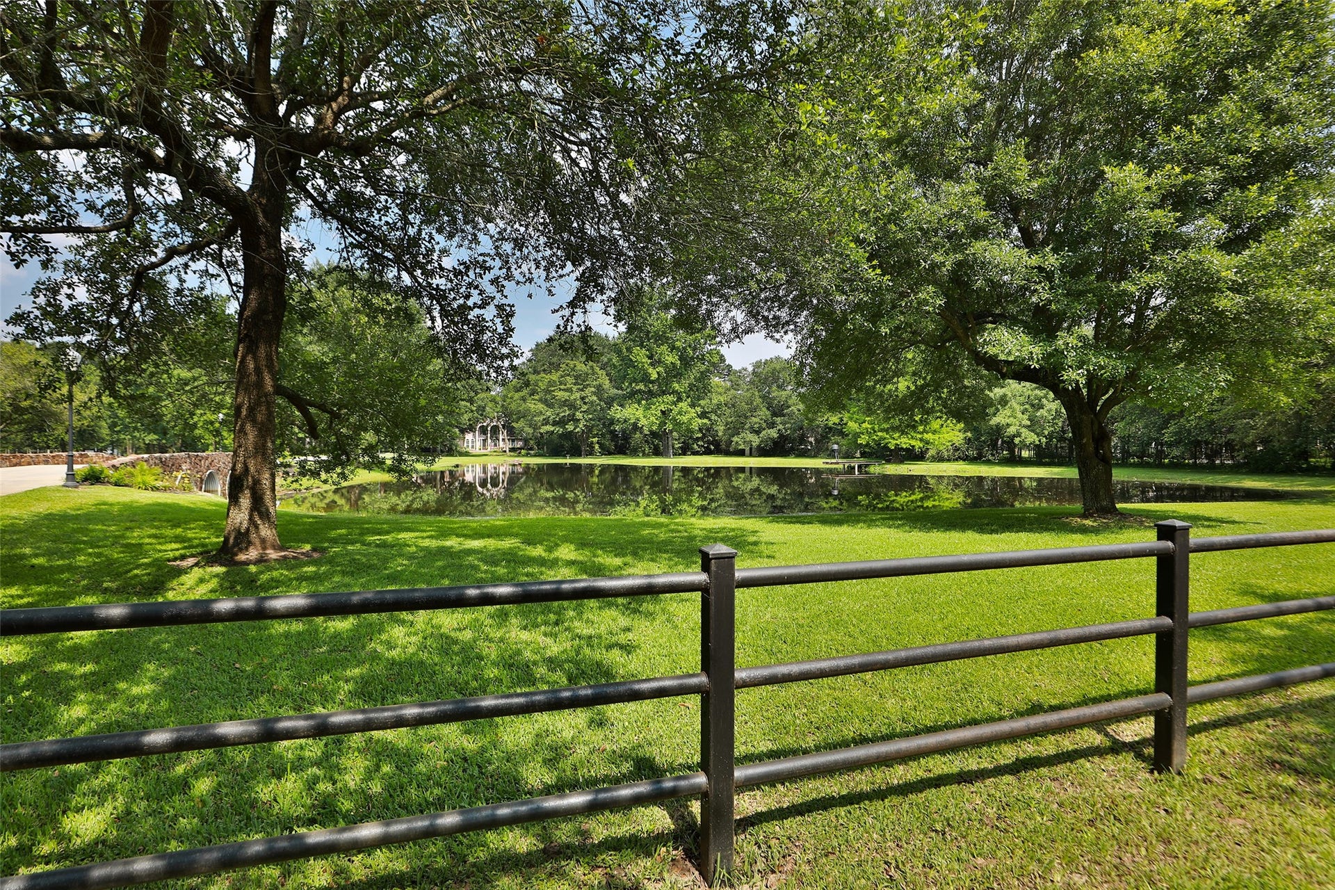 Framed by mature oak shade trees and a classic black pipe fence, this picturesque view showcases the estate's serene, stocked pond with the custom bridge creating a stunning focal point. The lush park-like setting offers approximately 2.47 acres of irrigated grounds supported by a sprinkler system, keeping the landscape vibrant year-round.