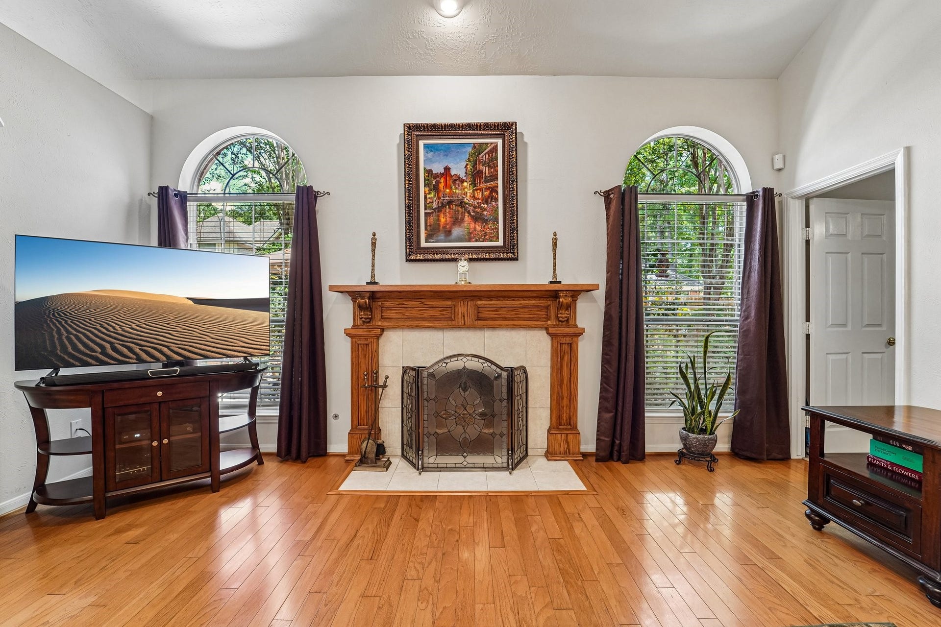 14 Serene Creek Place, The Woodlands, TX 77382 Living room features a classic fireplace with a wooden mantle, flanked by two arched windows that invite natural light. Warm hardwood floors and tasteful decor create a cozy yet sophisticated ambiance.