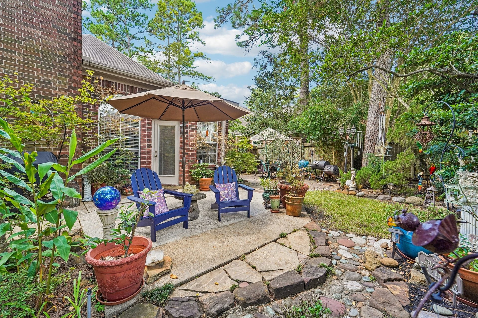 14 Serene Creek Place, The Woodlands, TX 77382 Charming backyard oasis with a cozy patio, featuring Adirondack chairs under a large umbrella, surrounded by lush greenery and decorative elements, perfect for relaxation.