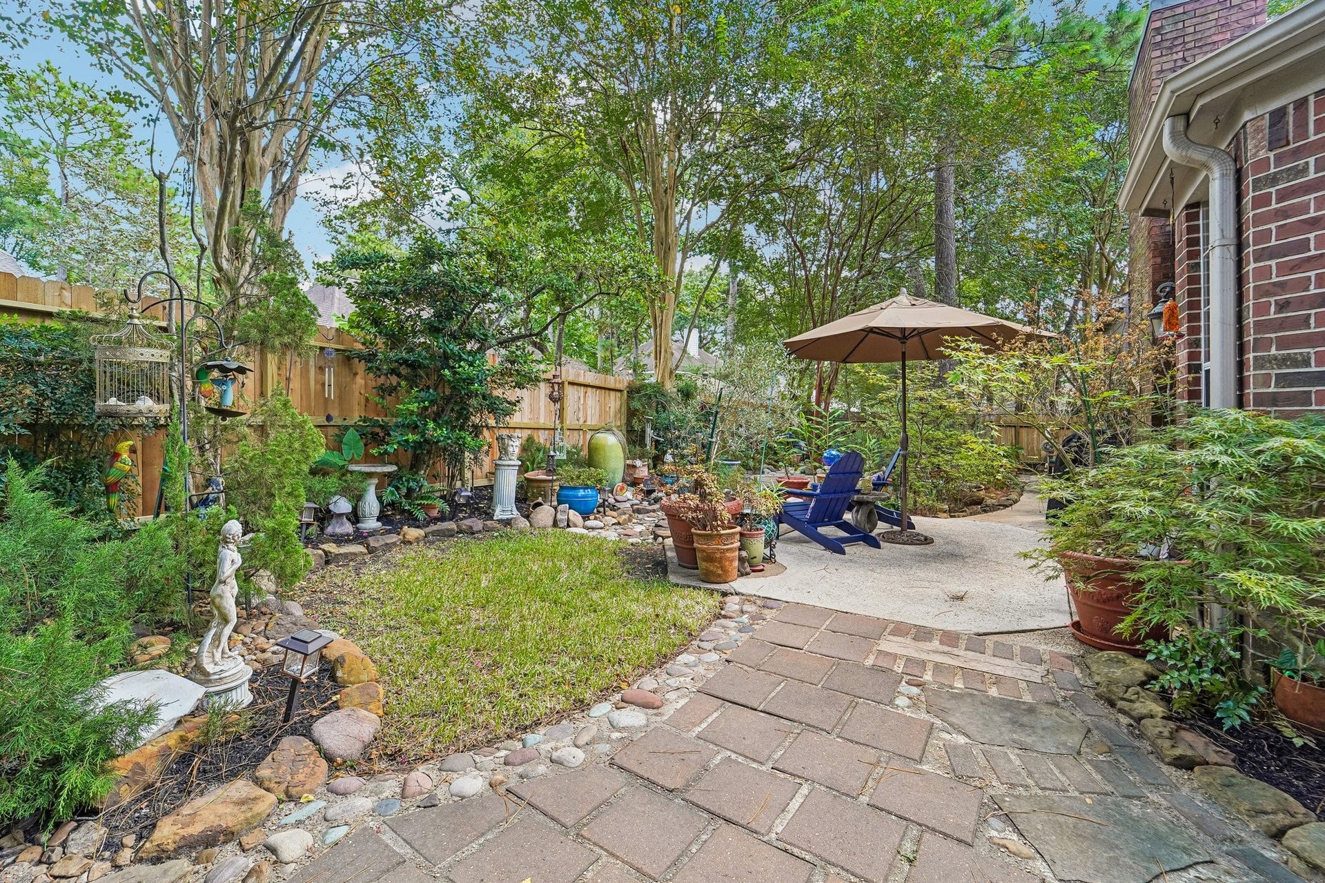 14 Serene Creek Place, The Woodlands, TX 77382 Charming backyard oasis with lush greenery, a cozy seating area under a large umbrella, and a delightful mix of potted plants and garden accents, perfect for relaxation and outdoor enjoyment.