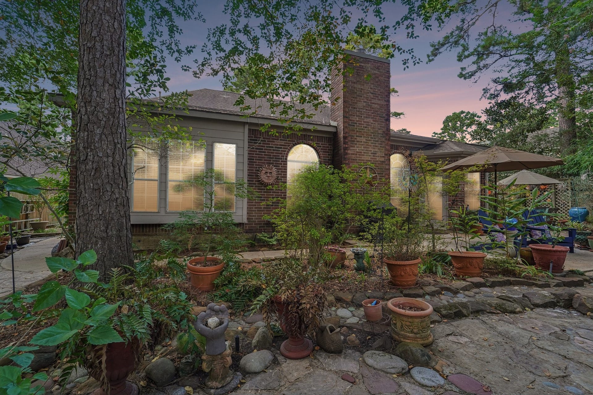 14 Serene Creek Place, The Woodlands, TX 77382 Charming brick home with large windows, nestled in a lush garden setting featuring mature trees, vibrant potted plants, and a cozy patio area perfect for outdoor relaxation.
