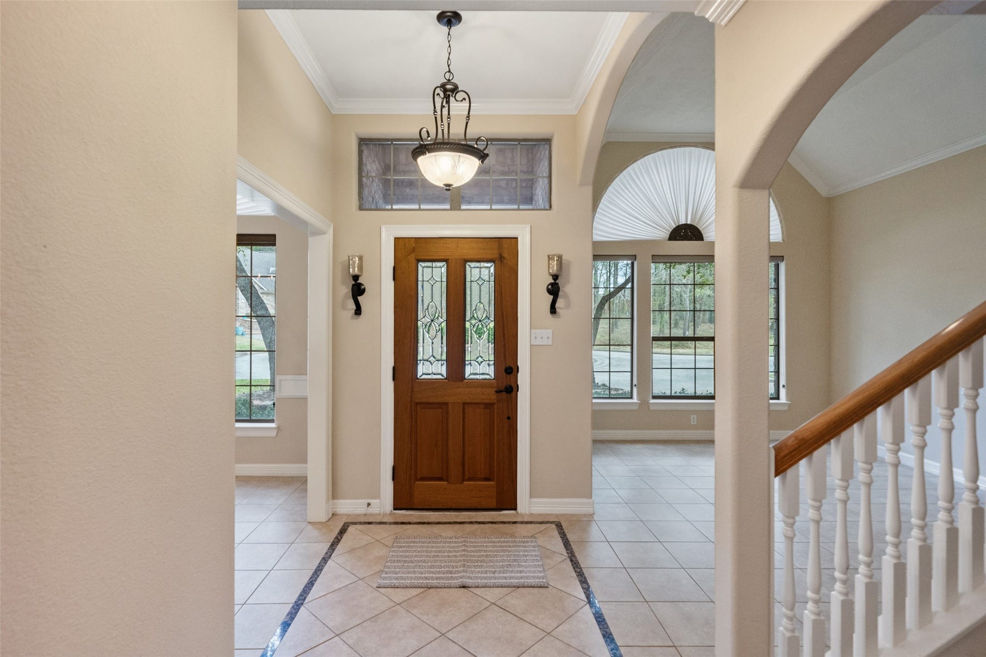 901 Longmire Road, #28, Conroe, TX 77304 This inviting entryway features a wooden front door with decorative glass panels, elegant tile flooring, and a chandelier. Large windows bring in natural light, and the space opens up to a staircase with white railings, offering a welcoming and spacious feel.