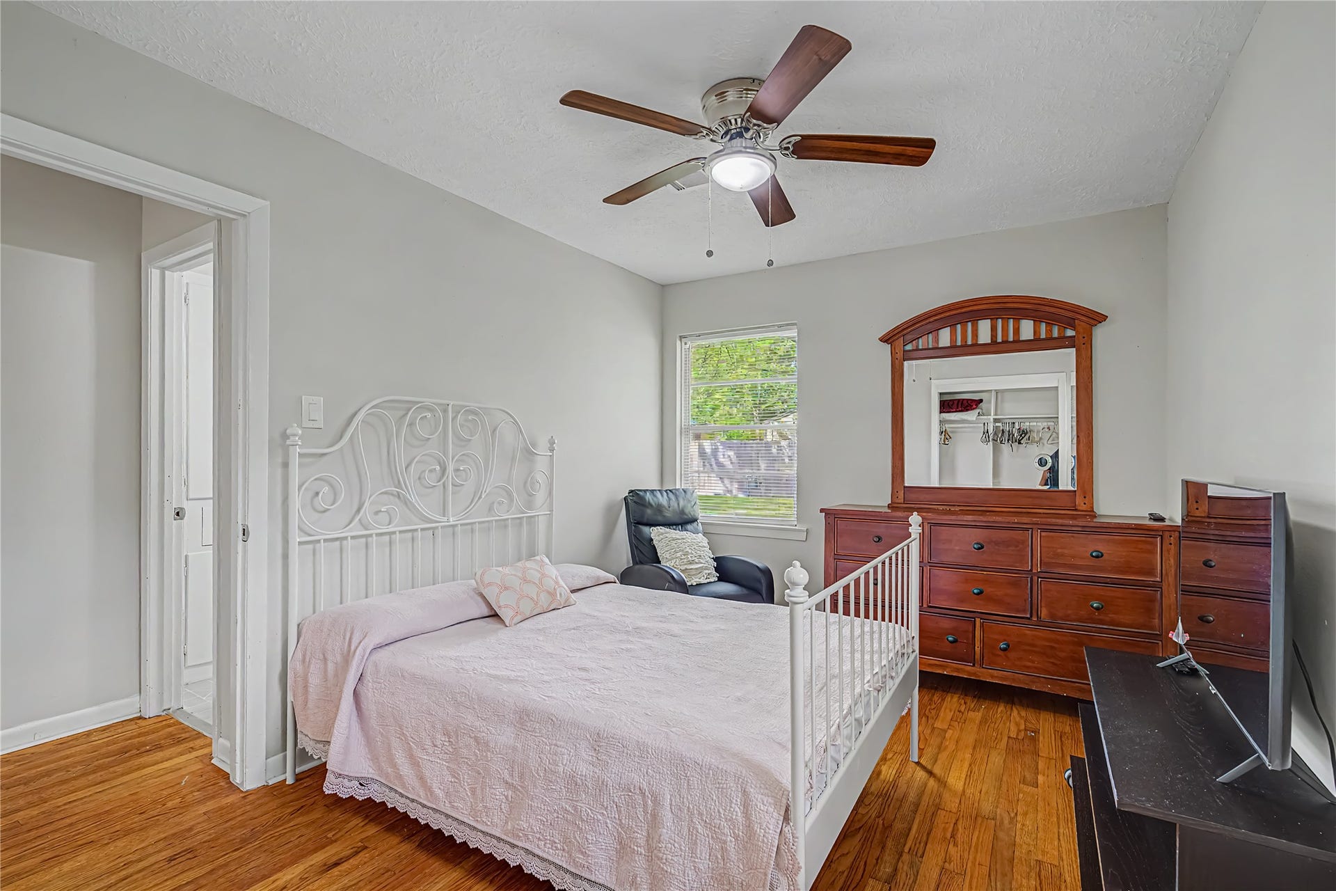Cozy bedroom featuring warm hardwood floors, and a rich wooden dresser, all complemented by ample natural light from the window.