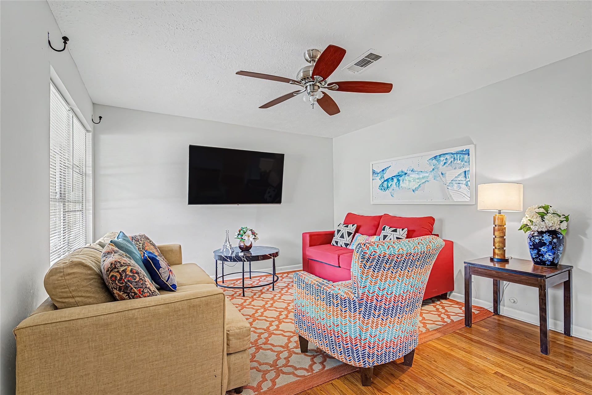 Bright and inviting living room featuring stylish seating, a modern ceiling fan, and a wall-mounted TV, complemented by vibrant accents and natural light.