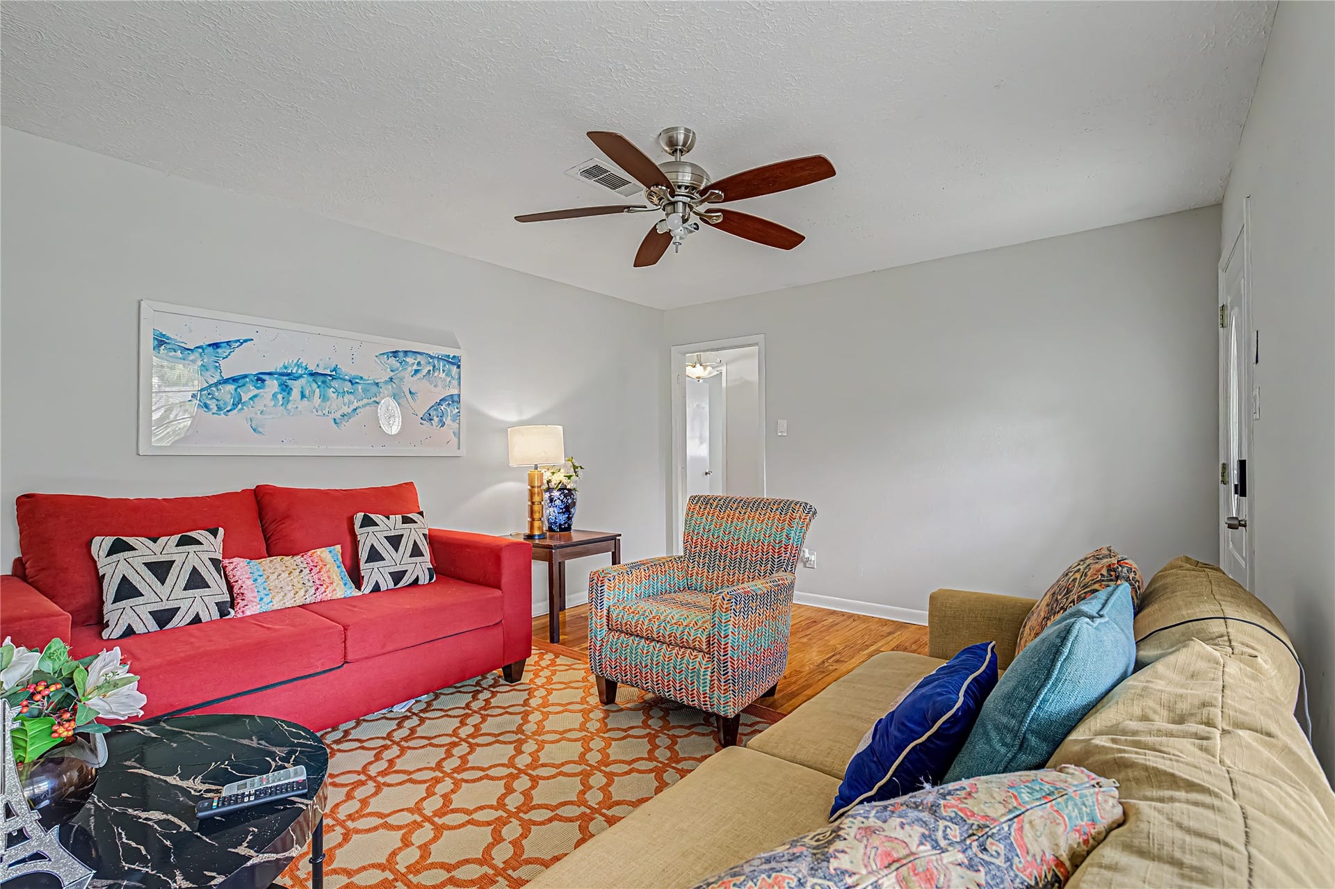 Charming living room with vibrant furnishings, featuring a bold red couch and colorful accent chair, enhanced by a ceiling fan and warm lighting.