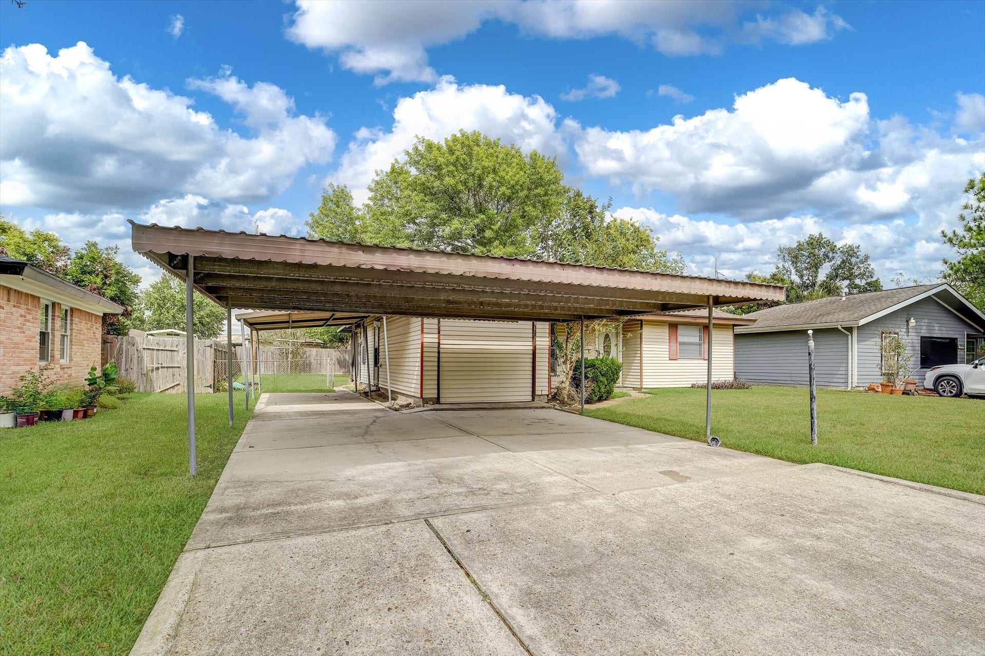Spacious driveway with a large covered carport, perfect for multiple vehicles, set against a backdrop of lush greenery and a fenced backyard.