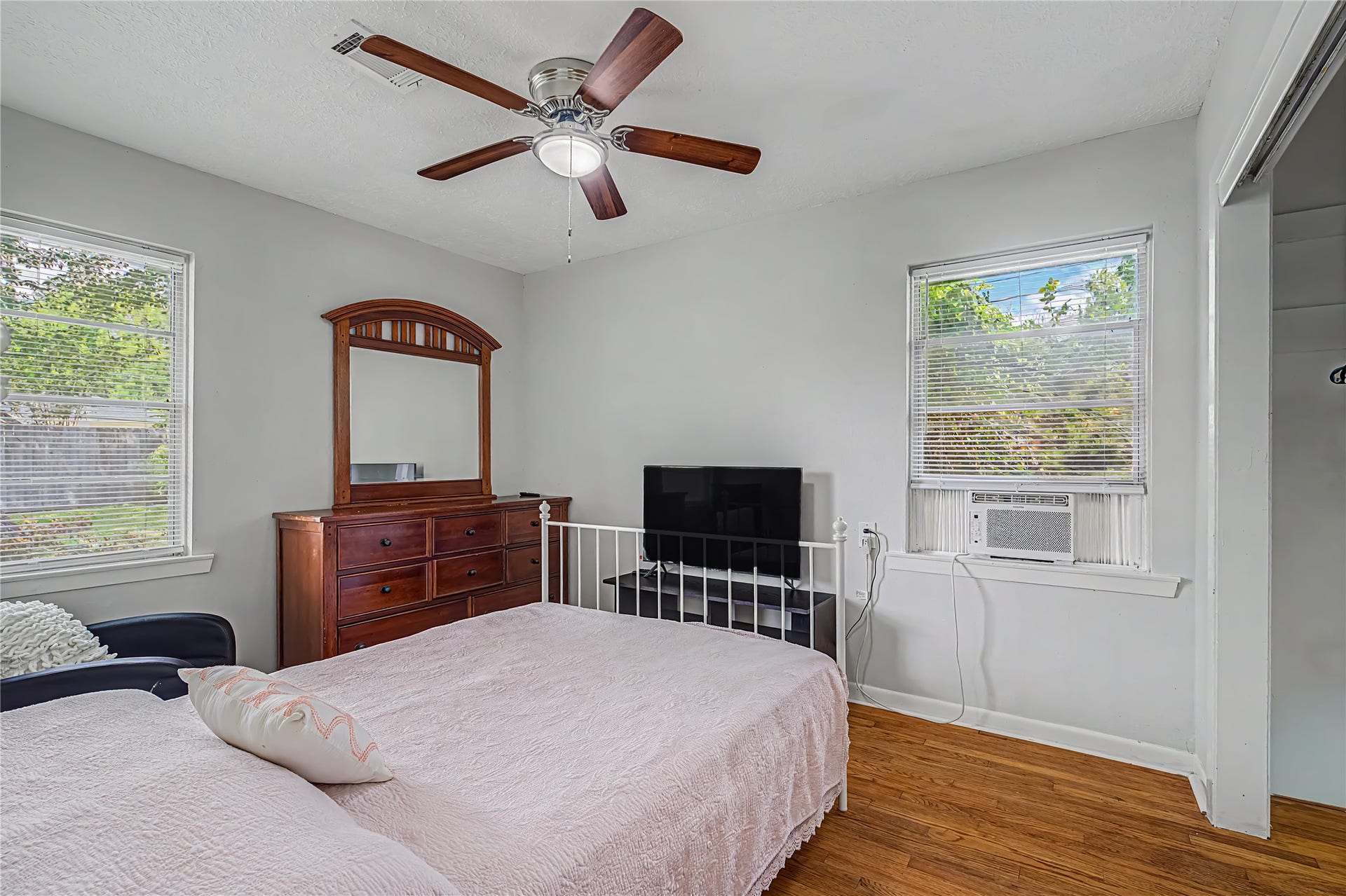 Cozy bedroom with natural light, featuring a ceiling fan, hardwood floors, and a spacious dresser with mirror.