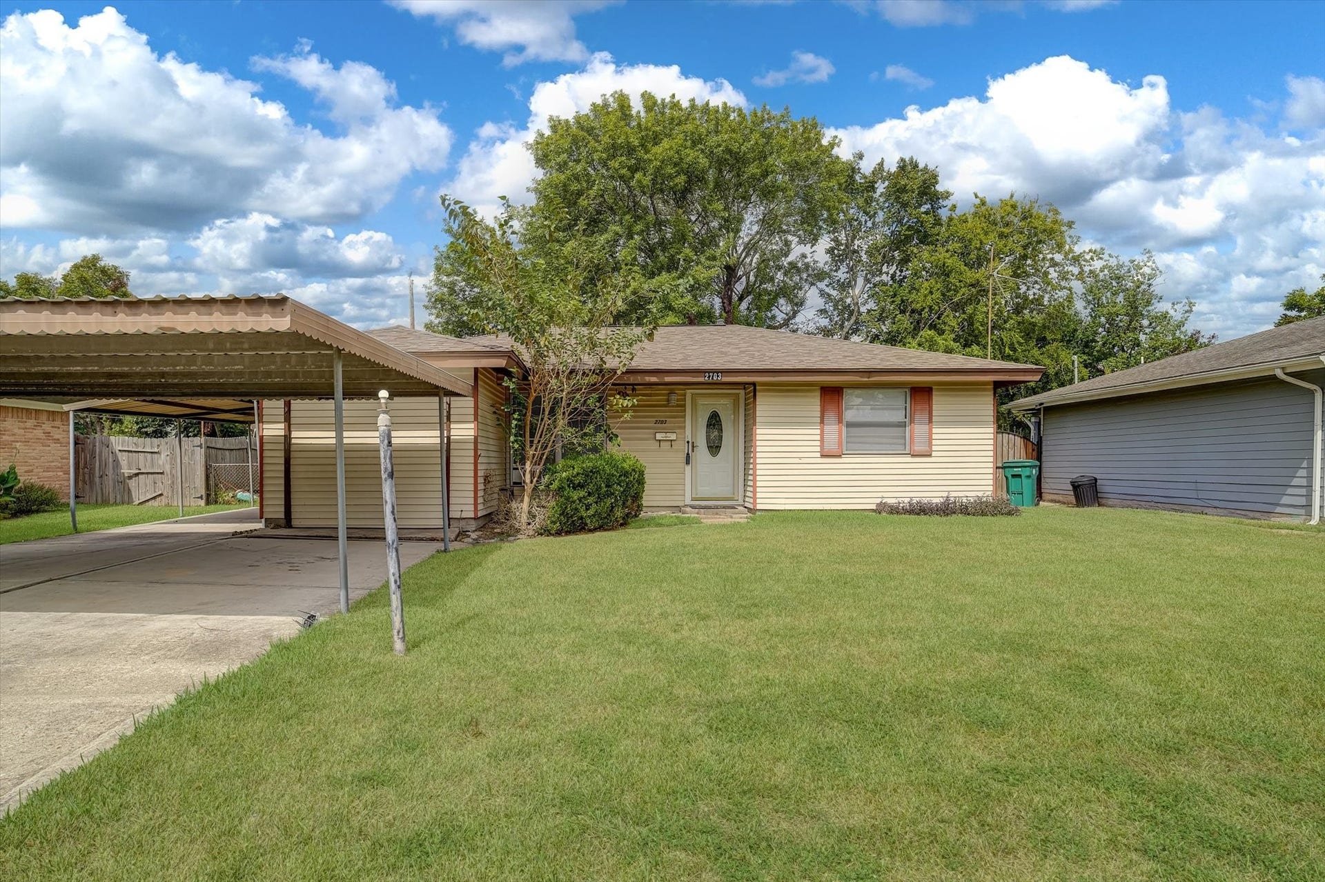 Charming single-story home with a welcoming front yard and covered carport, set against a backdrop of mature trees.