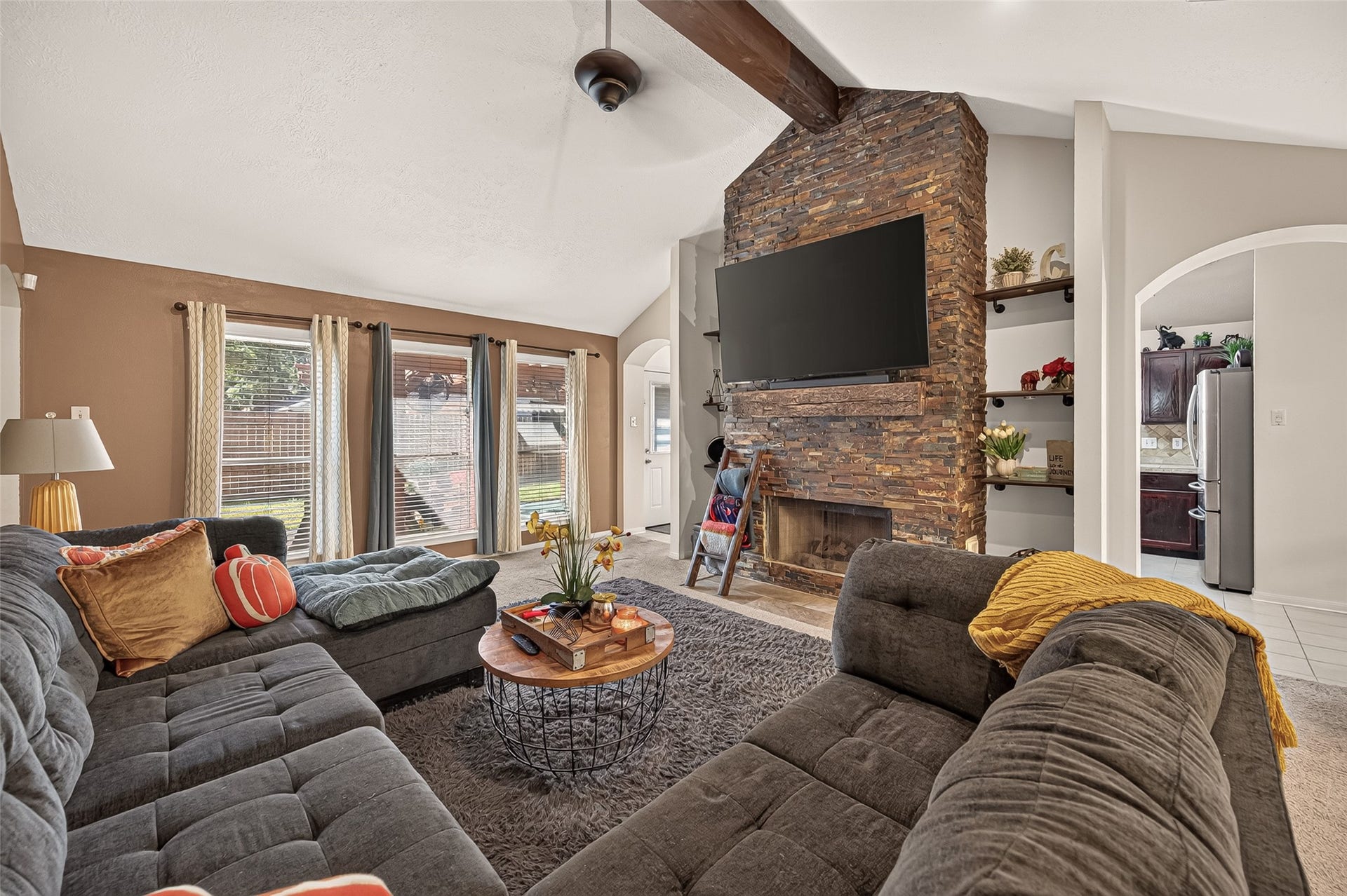 1615 Glacier Blue Drive, Fresno, TX 77545 Spacious living room featuring a vaulted ceiling with a rustic beam, a striking stone fireplace, and large windows that fill the space with natural light.