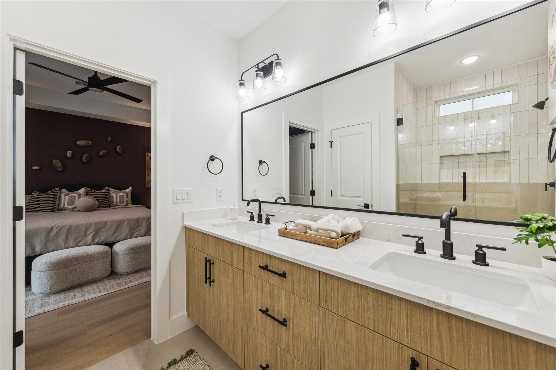 Elegant bathroom with a double vanity featuring sleek black fixtures and a large mirror, seamlessly connecting to a cozy bedroom through a sliding door.